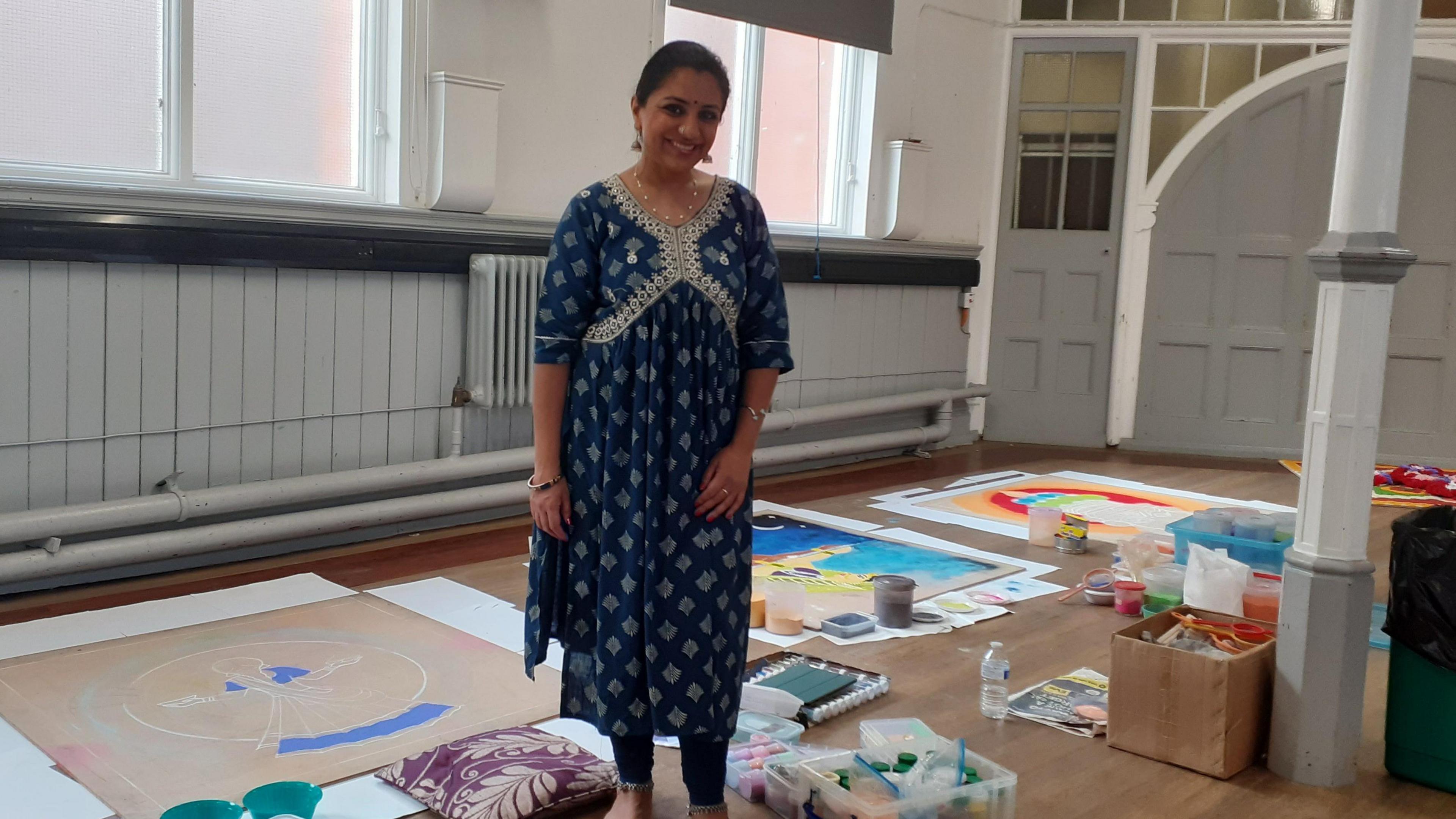 A woman smiling in front of large artworks being created inside a community centre hall