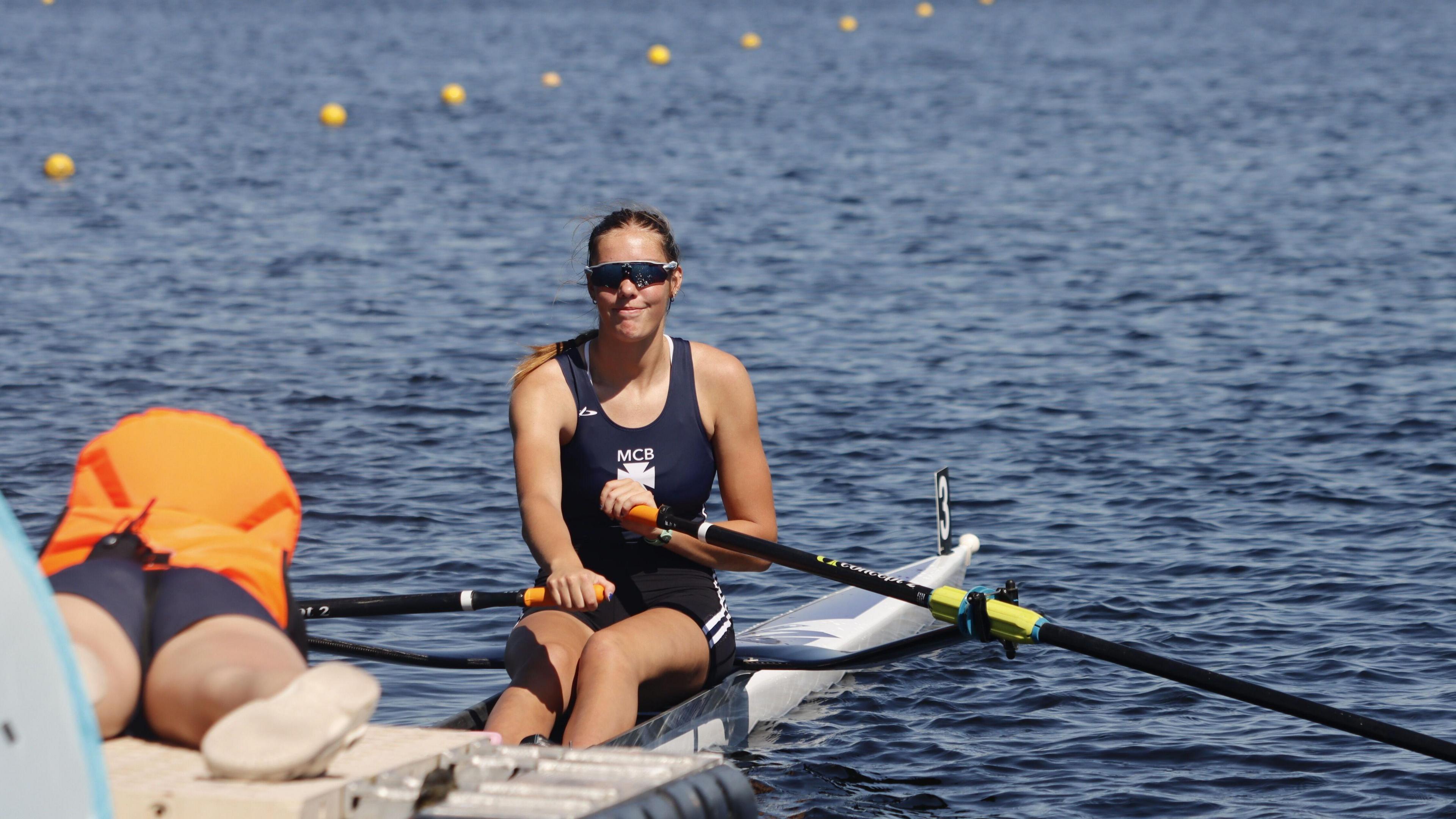 Sophia sitting in her white row boat, holding two black oars. Sophia is smiling whilst sitting in a row boat in a blue lake, wearing her Methodist row club grey swimwear along with a pair of sun visors.
