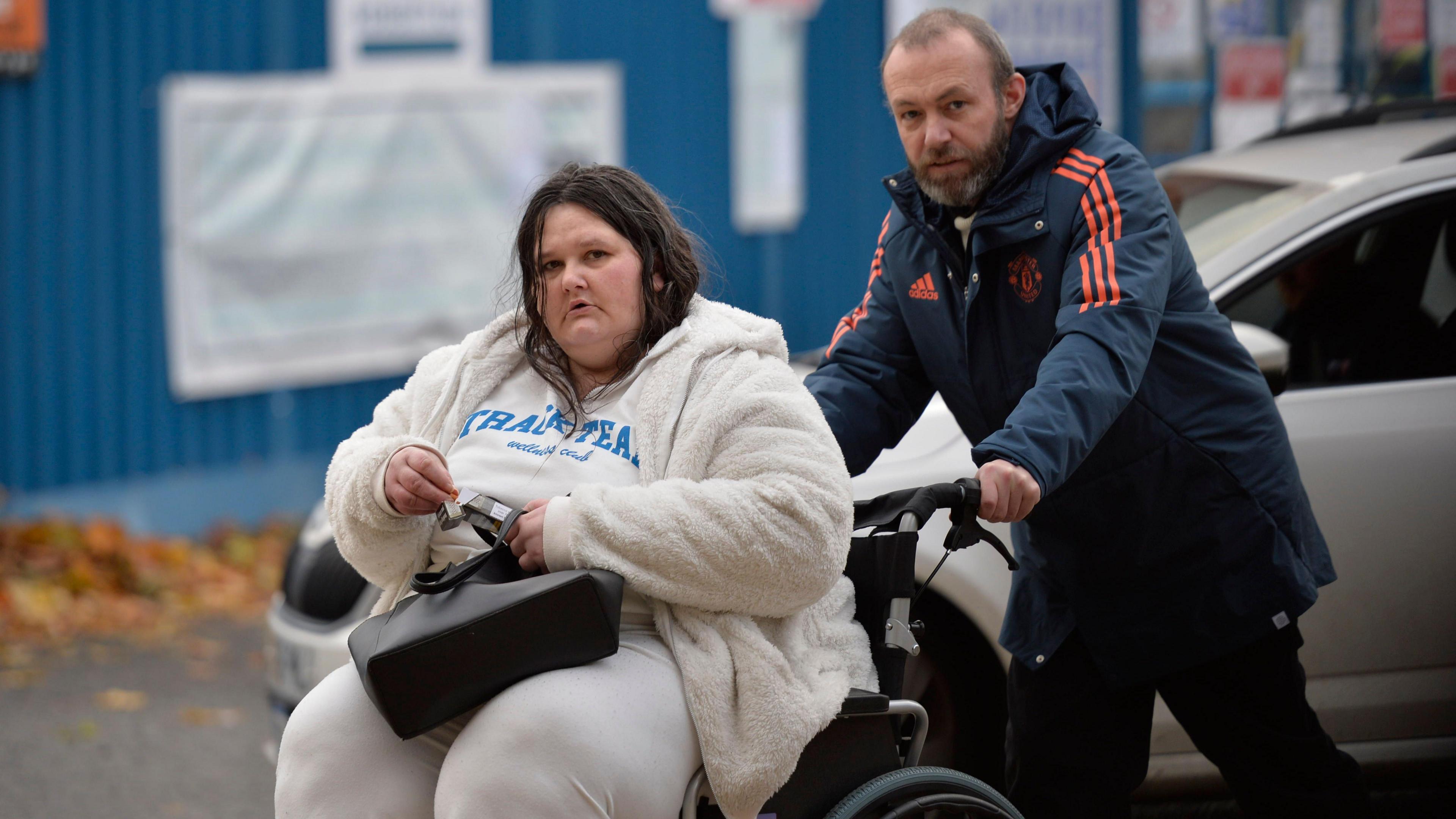 Paul Brannan photographed while pushing Lesley Williams in a wheelchair outside the High Court in Glasgow. Williams has long, dark hair with a middle parting and is wearing a white fleece, white t-shirt with blue writings and white trousers. She is holding a black handbag, which is balanced on her lap, and is looking straight at the camera with a serious expression. Brannan is leaning forward  pushing the wheelchair. He is wearing black trousers and a navy blue Manchester United jacket with the club crest, adidas logo and three stripes on the arms in red. Brannan has dark thinning hair and a greying beard. He is also looking at the camera with a serious expression.