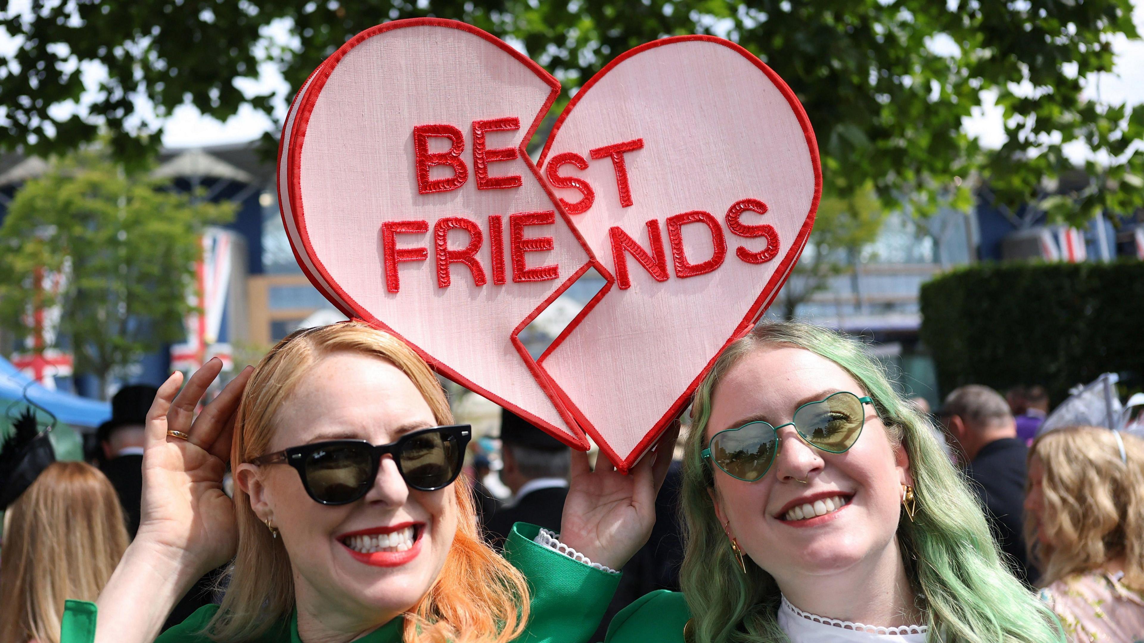 Two women wearing fascinators which together spell "best friends" in the style of a Love Heart sweet. 