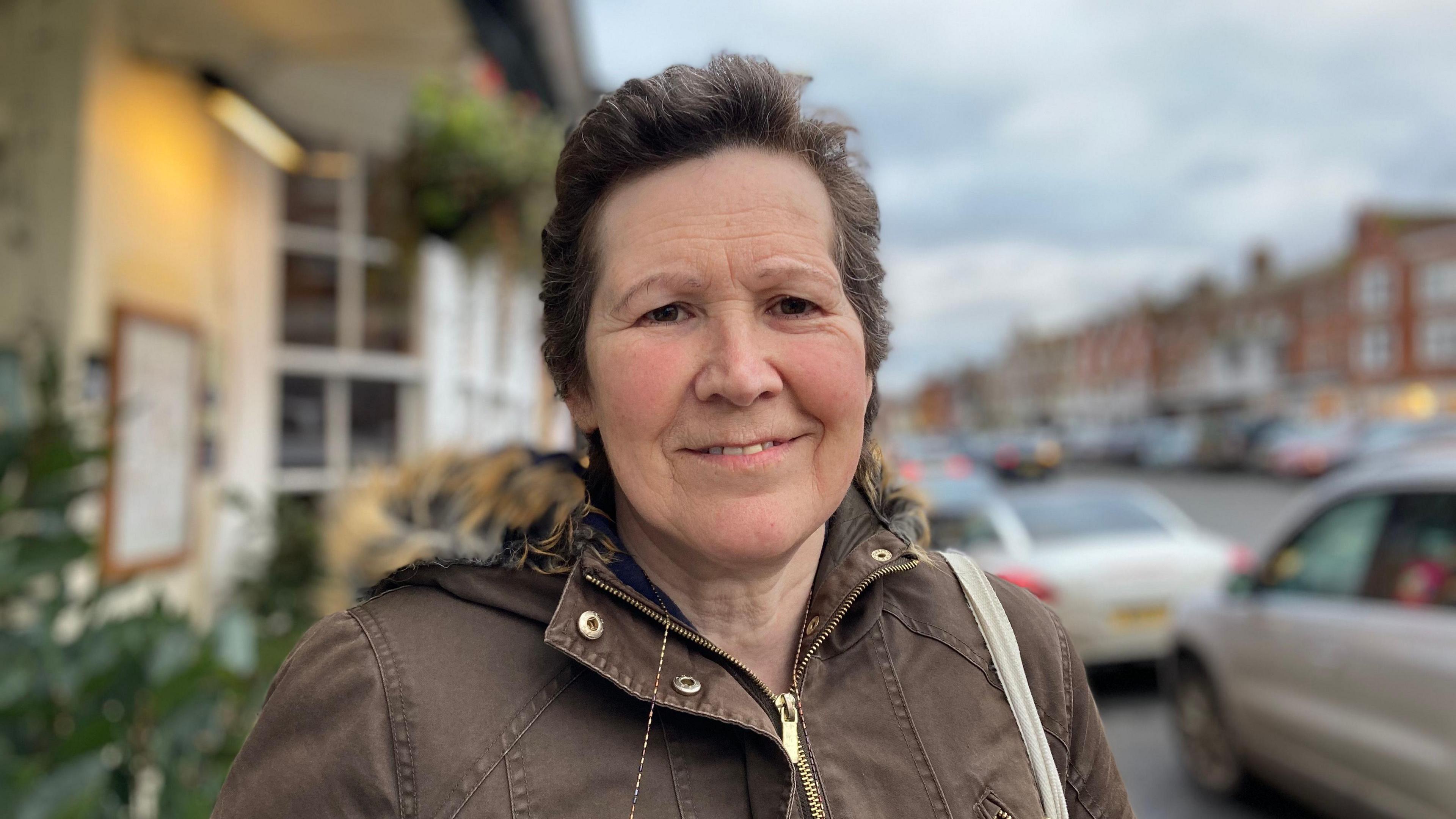 Jackie looks into the camera wearing a brown winter coat standing by shops in the High Street