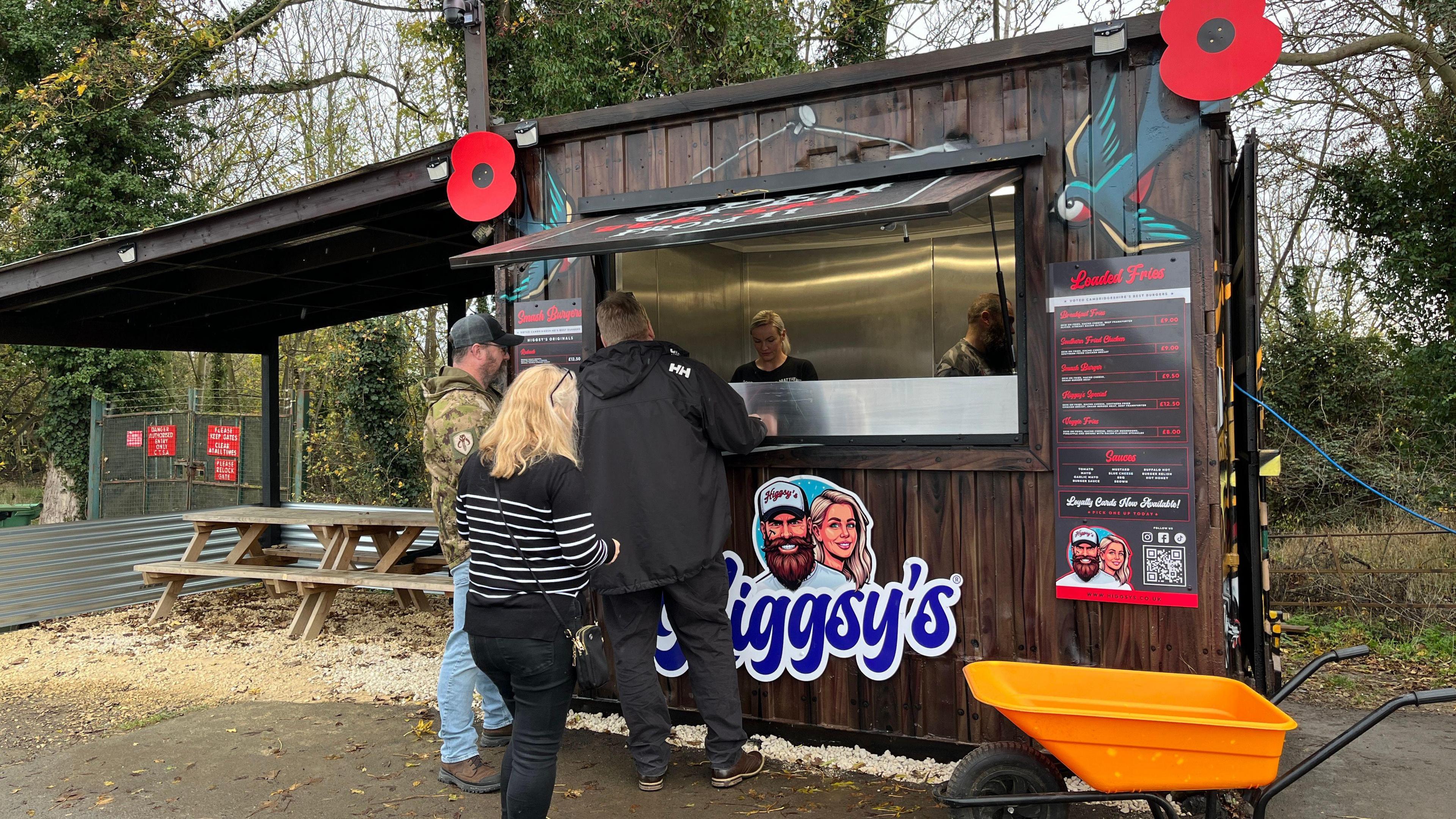 A dark brown wooden burger shed with a blue Higgsy's sign on it. To the left of the shed, covered by a roof, here is a seating area with two picnic tables. Amber, a woman in a black shirt, is serving customers from inside. Adam can be seen preparing burgers. Three customers are queuing at the serving hatch.
