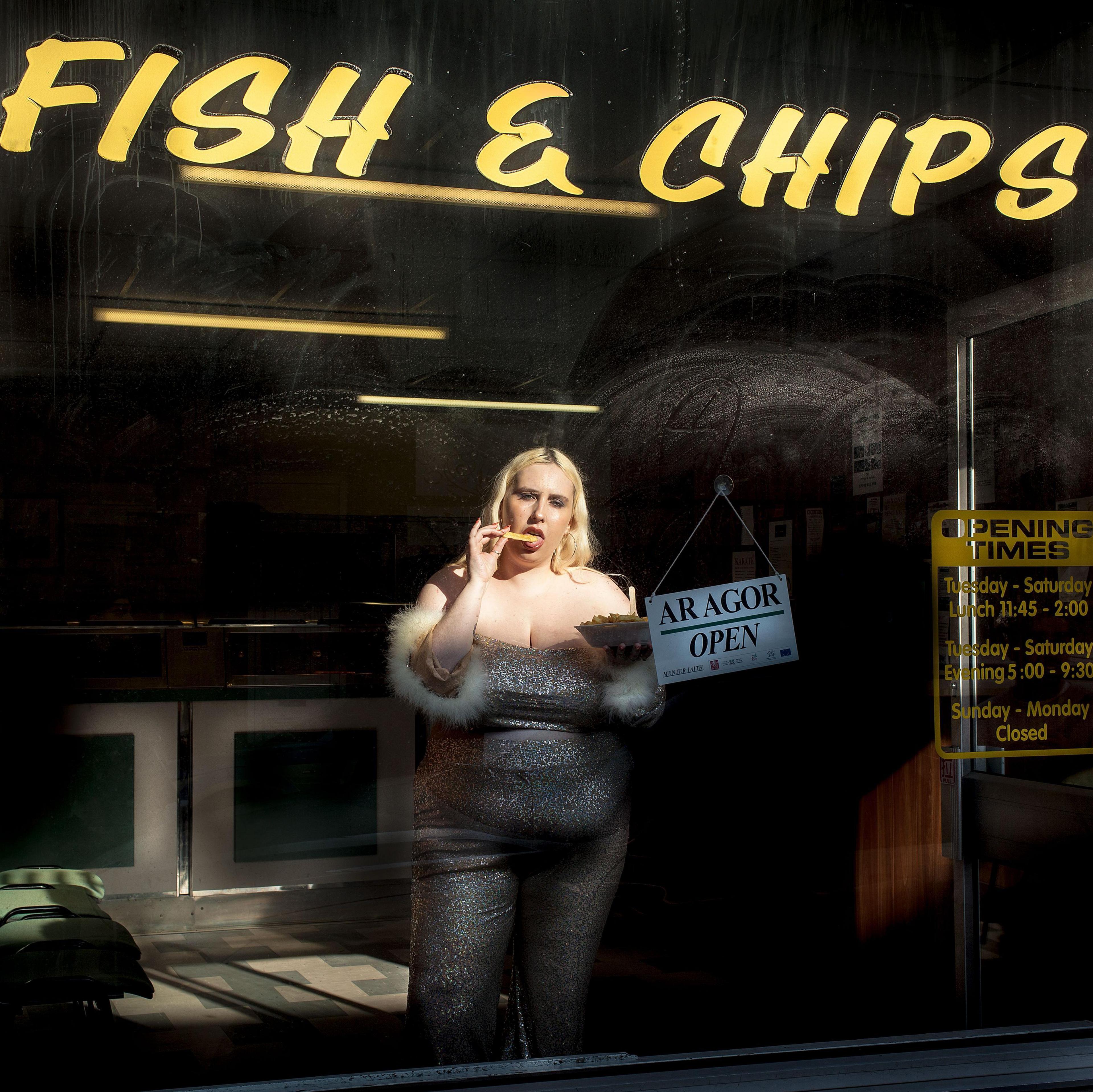 Shot from outside a chip shop, Gina Tonic is wearing a silver strapless jumpsuit and is eating chips