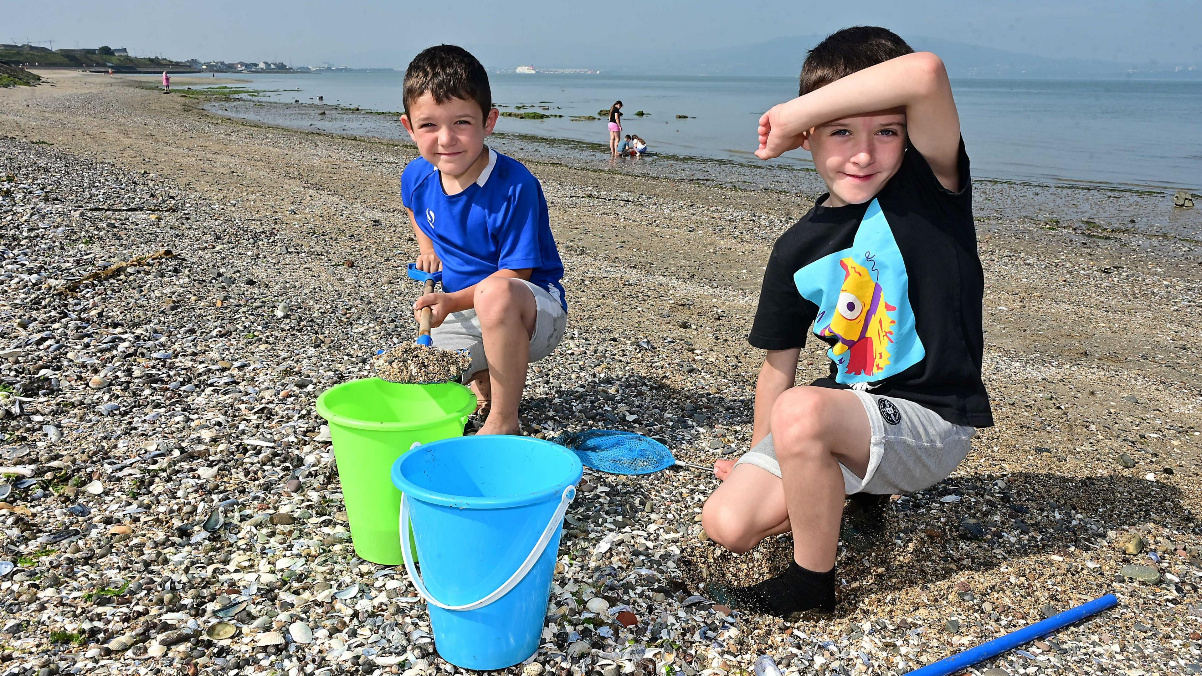 Two boys on a beach. They are both in shorts and T-shirts and have dark hair. There are two buckets in front of them - a green and blue one. A fishing net is also beside one of them.