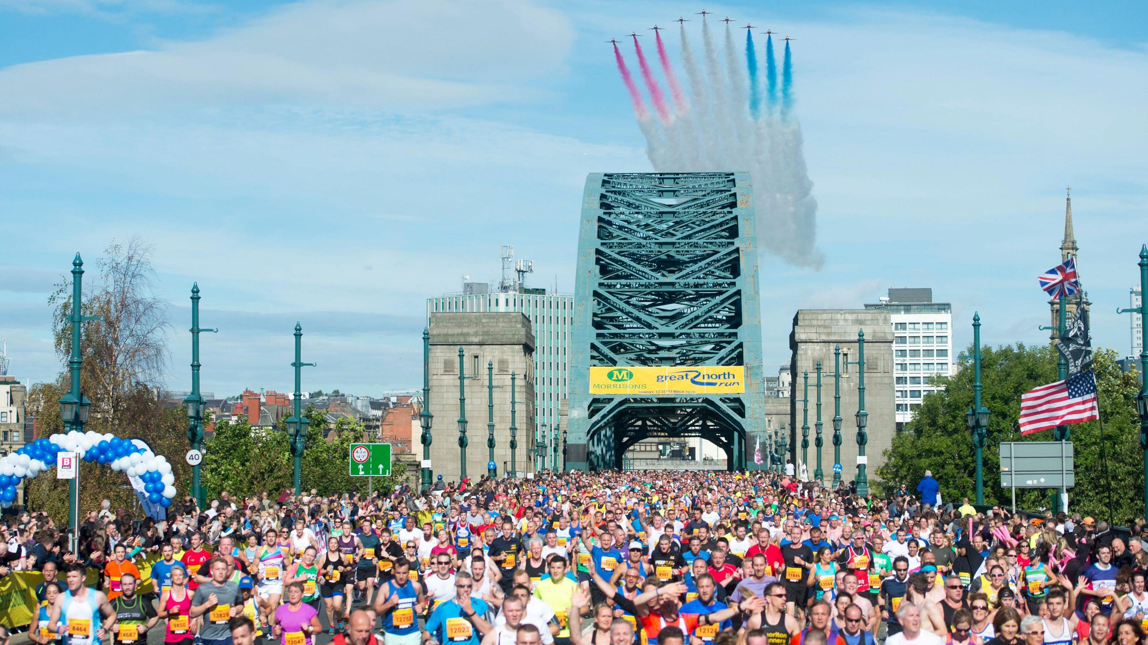 Crowds of runners, wearing colourful tops, run across the Tyne Bridge. A set of planes (Red Arrows) can been in the blue sky, leaving a trail of white, red and blue markings in the sky.