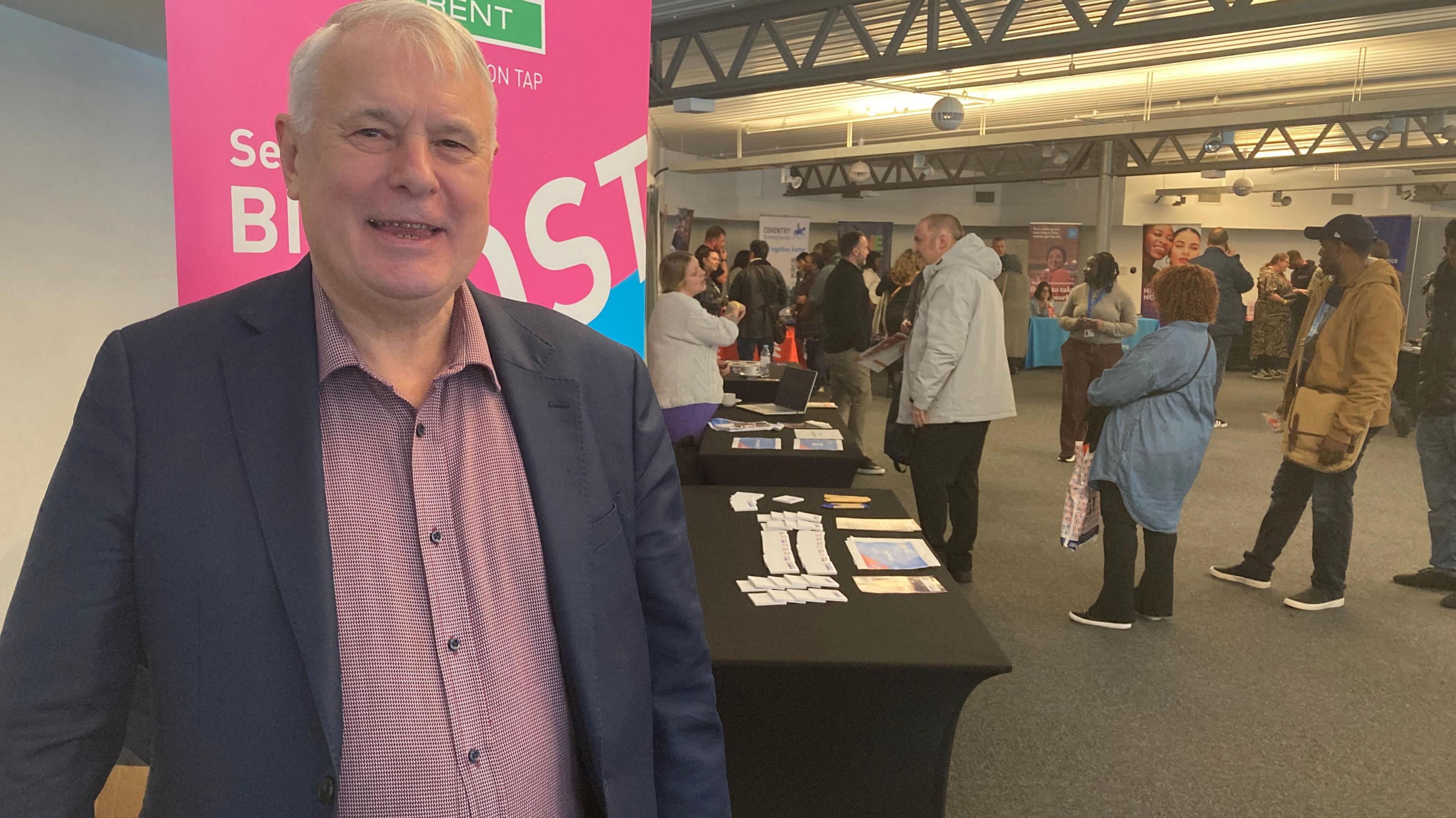 A man wearing a pink shirt and suit jacket stands in front of a big pink sign and black desks. The sign has 'Severn Trent' written at the top and people can be seen stood in the background.