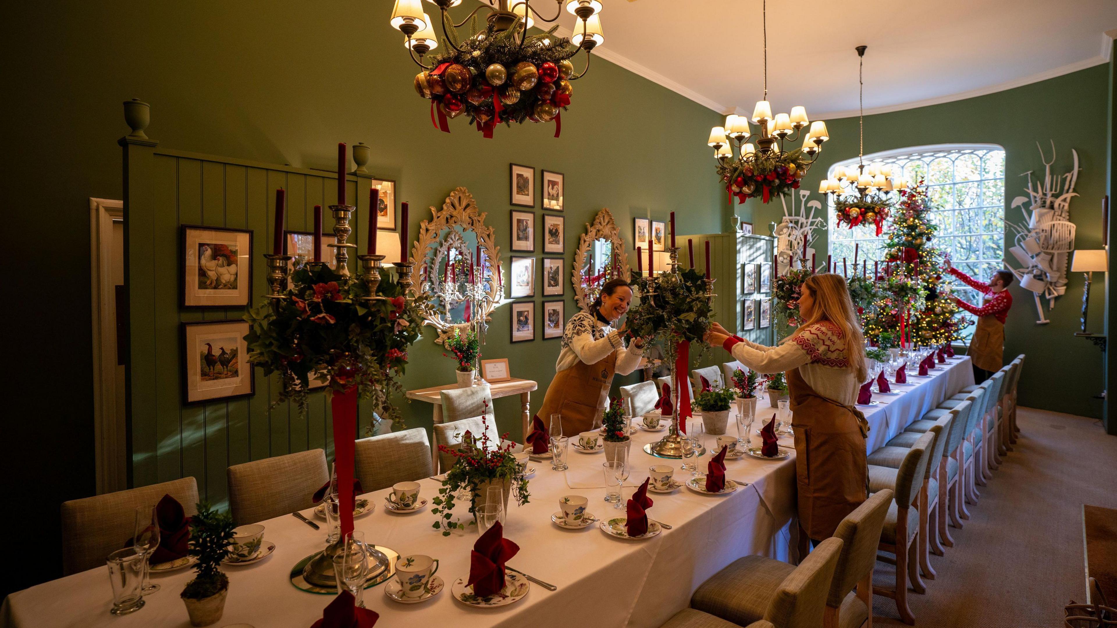 Staff decorate a long banqueting-style table at Highgrove. The table is laid with multiple places with ornate plates and red napkins, and the light fittings above it have festive wreaths around them. The chairs along the table are beige-coloured and the walls of the room are dark green