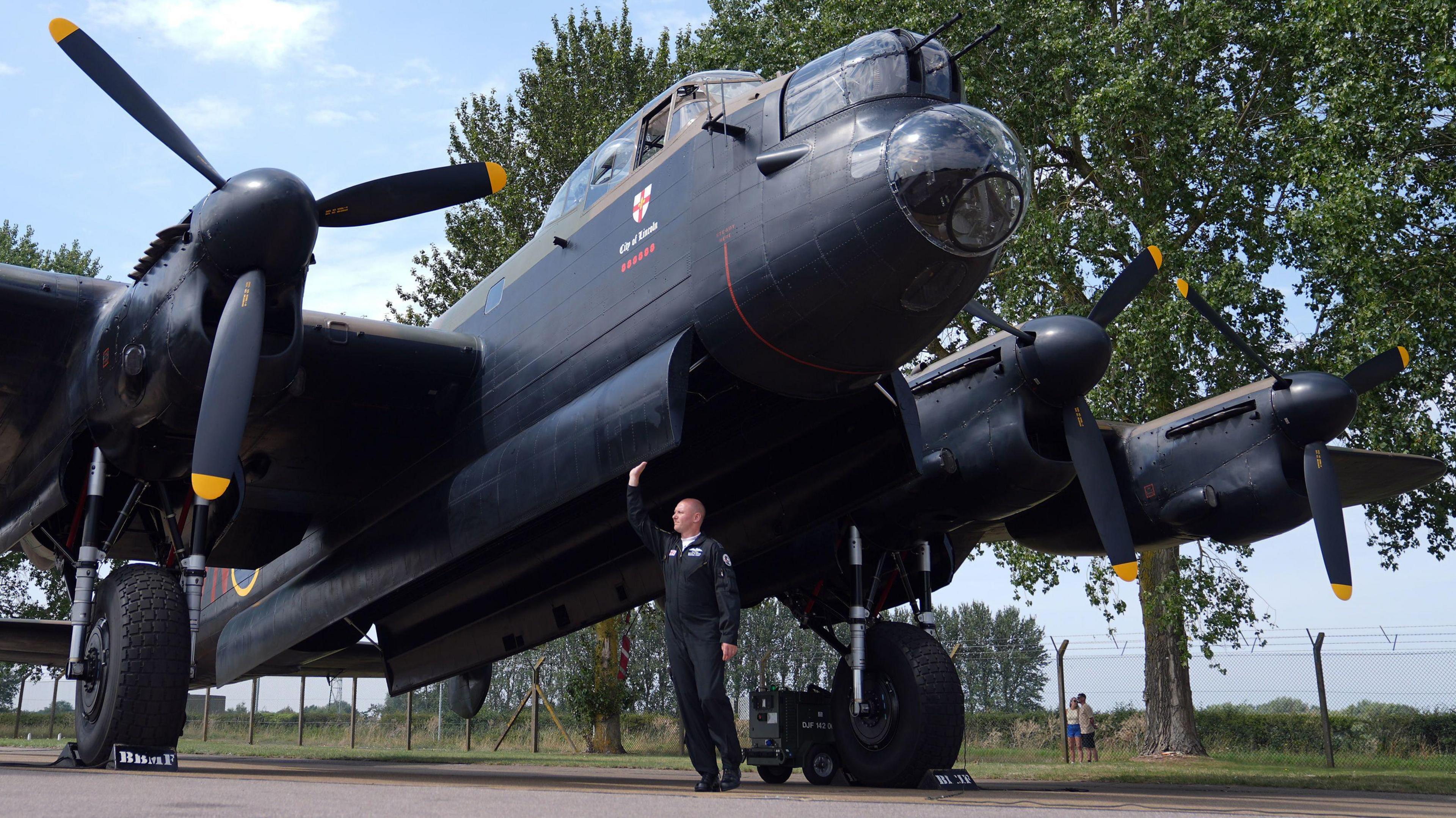 A close up picture taken from ground level of the Avro Lancaster PA474 which is stationary at RAF Coningsby in Lincolnshire. The underneath of the plane and large propellers at in the picture. Standing underneath the aircraft is the pilot Squadron Leader, Paul "Ernie" Wise, who is inspecting the plane.