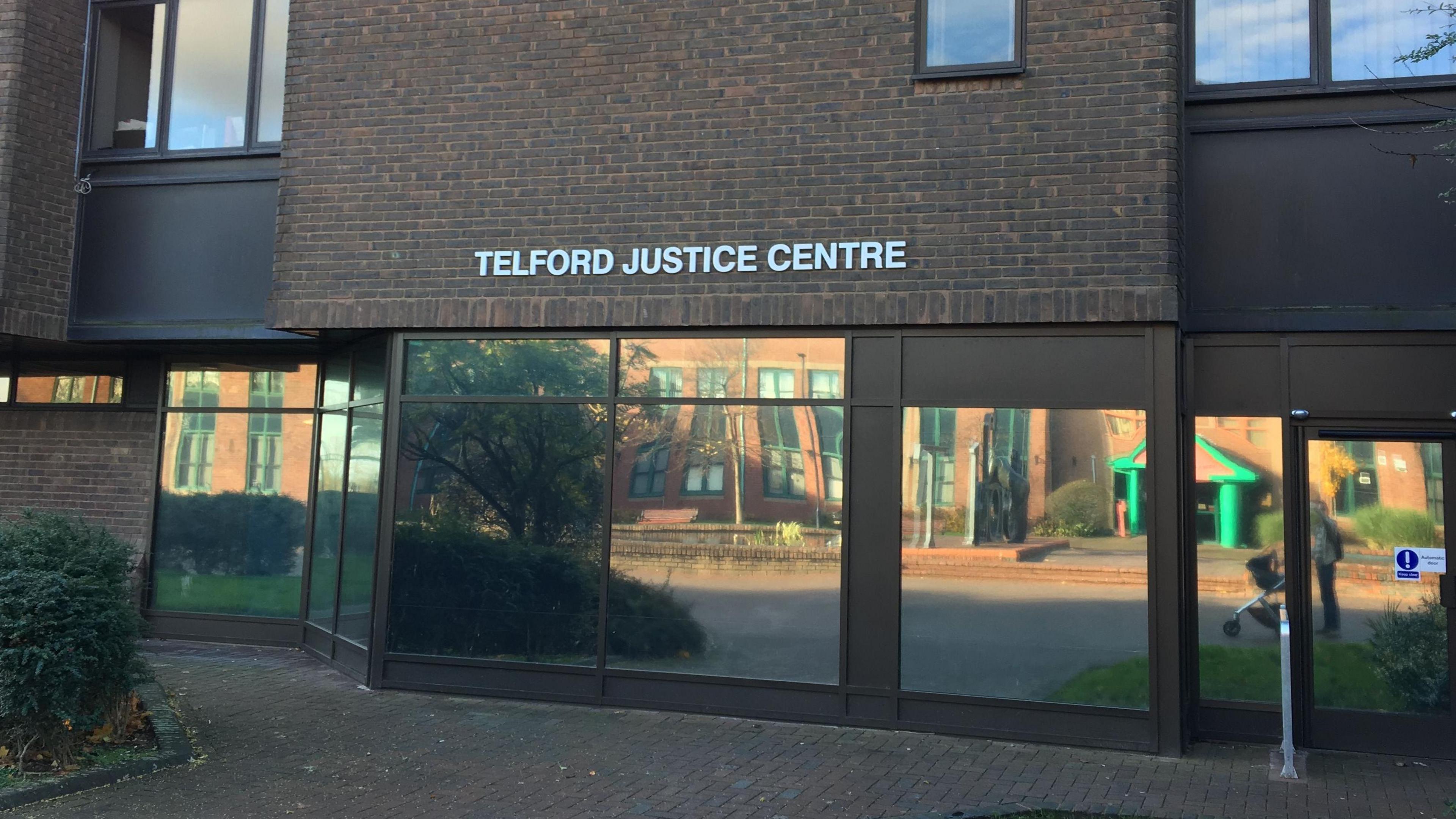 A nondescript looking court building. It is brick and brown and we can see reflective windows and an automatic door. A woman with a pushchair can be seen in the image. The words Telford Justice Centre are on the front of the building.