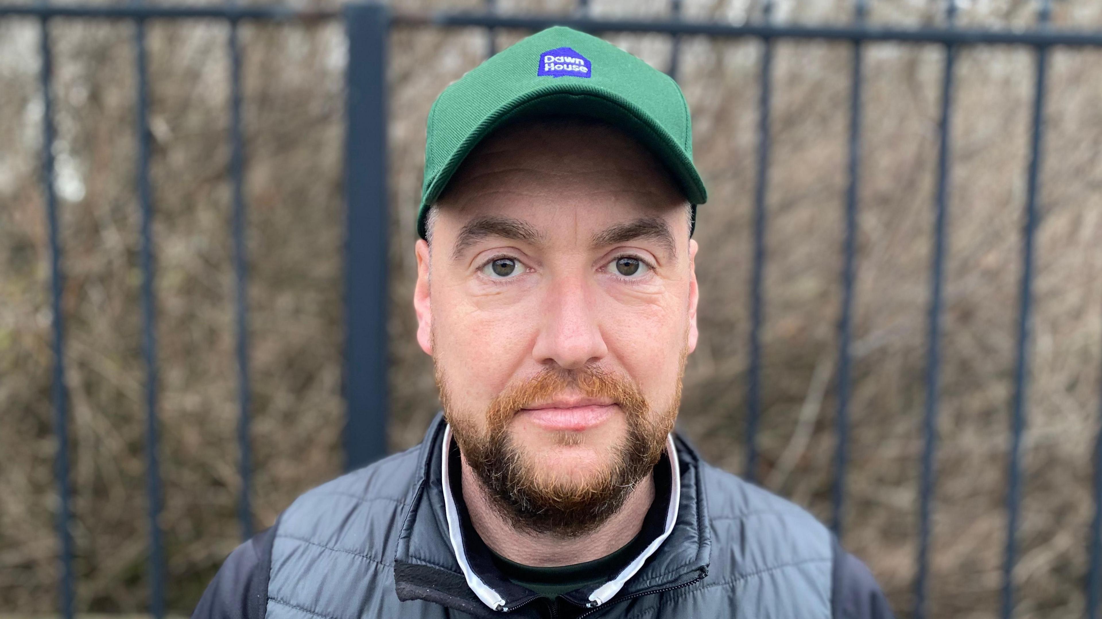 Glen McPherson, a man wearing a green cap and a black gilet with a short, dark brown beard.  