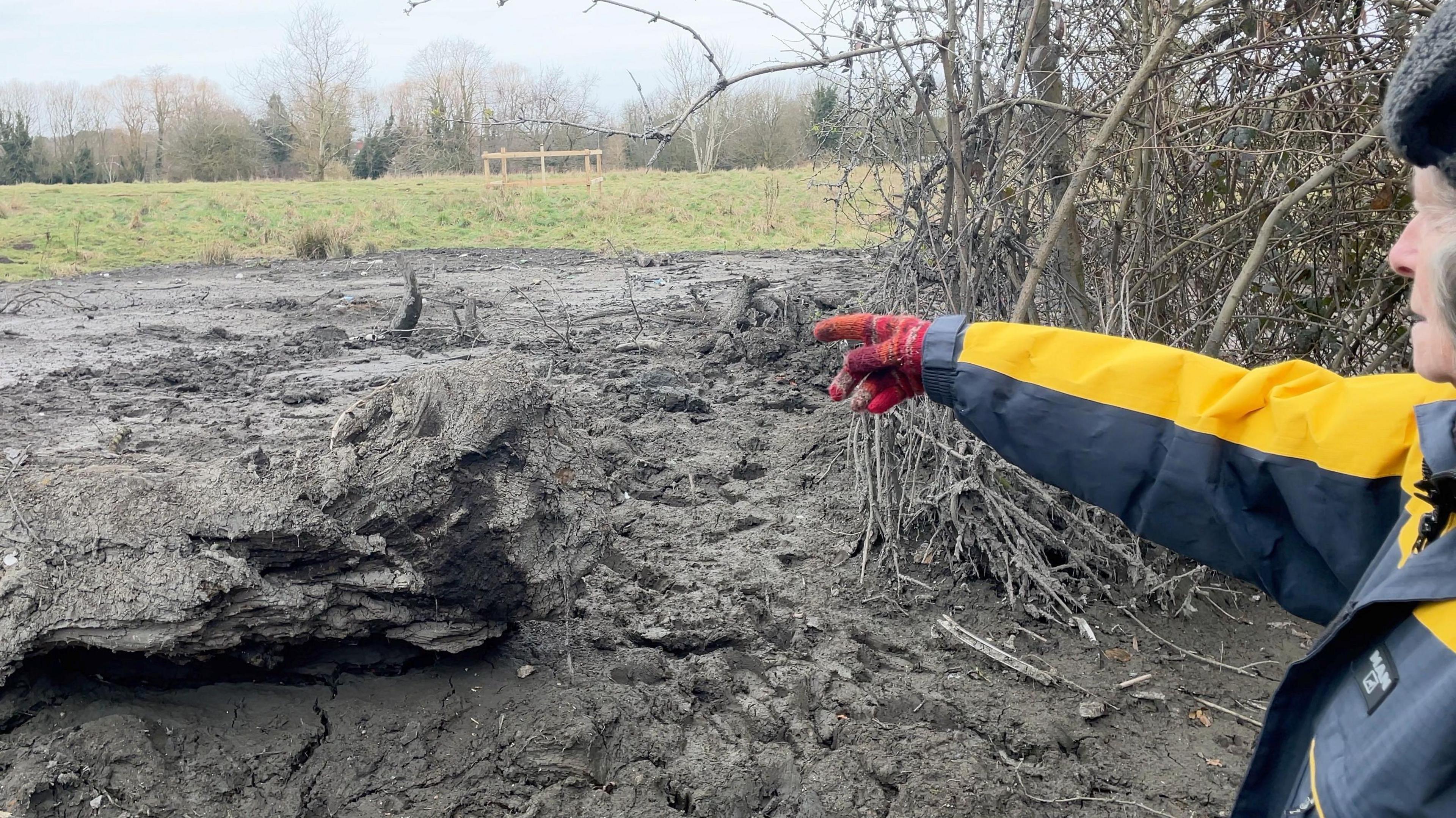 Gill in a yellow coat and warm hat points out the area she became stuck in.
You can see a large log to the left, and a dark-grey muddy area in front of her.