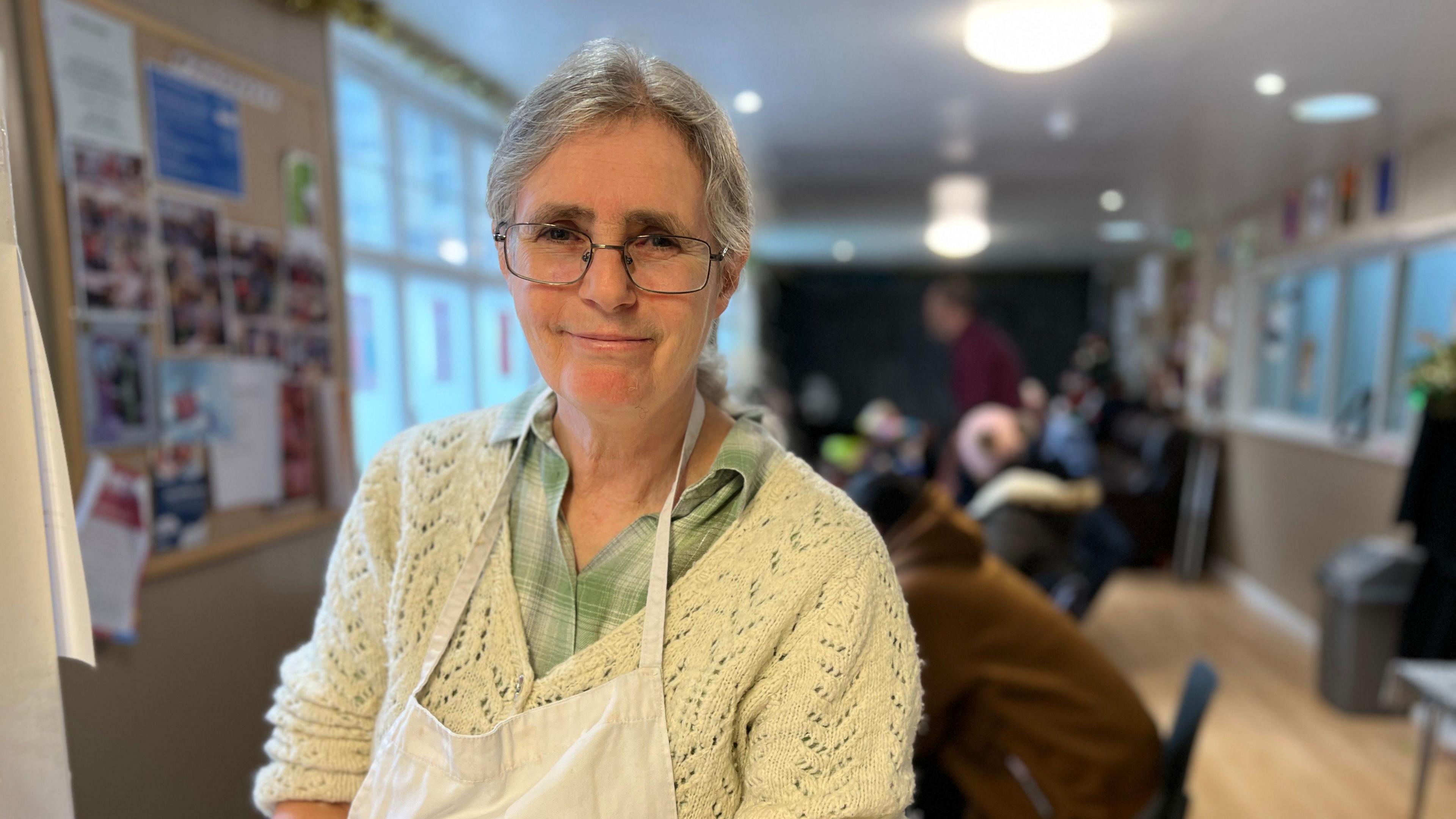 Juley Howard wearing a cream knit cardigan with a light green shirt underneath and an apron over the top. She is wearing glasses and is smiling. There is a room with people sitting at a table behind her.