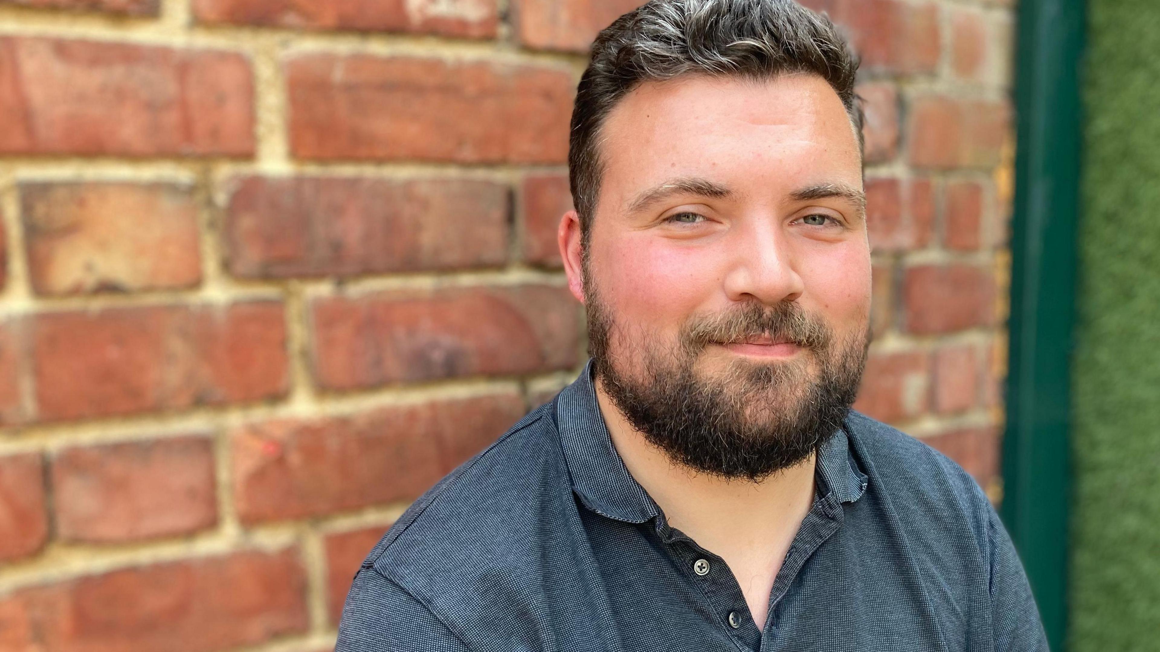 Man in a blue polo shirt looking at the camera and smiling with a brick wall behind him