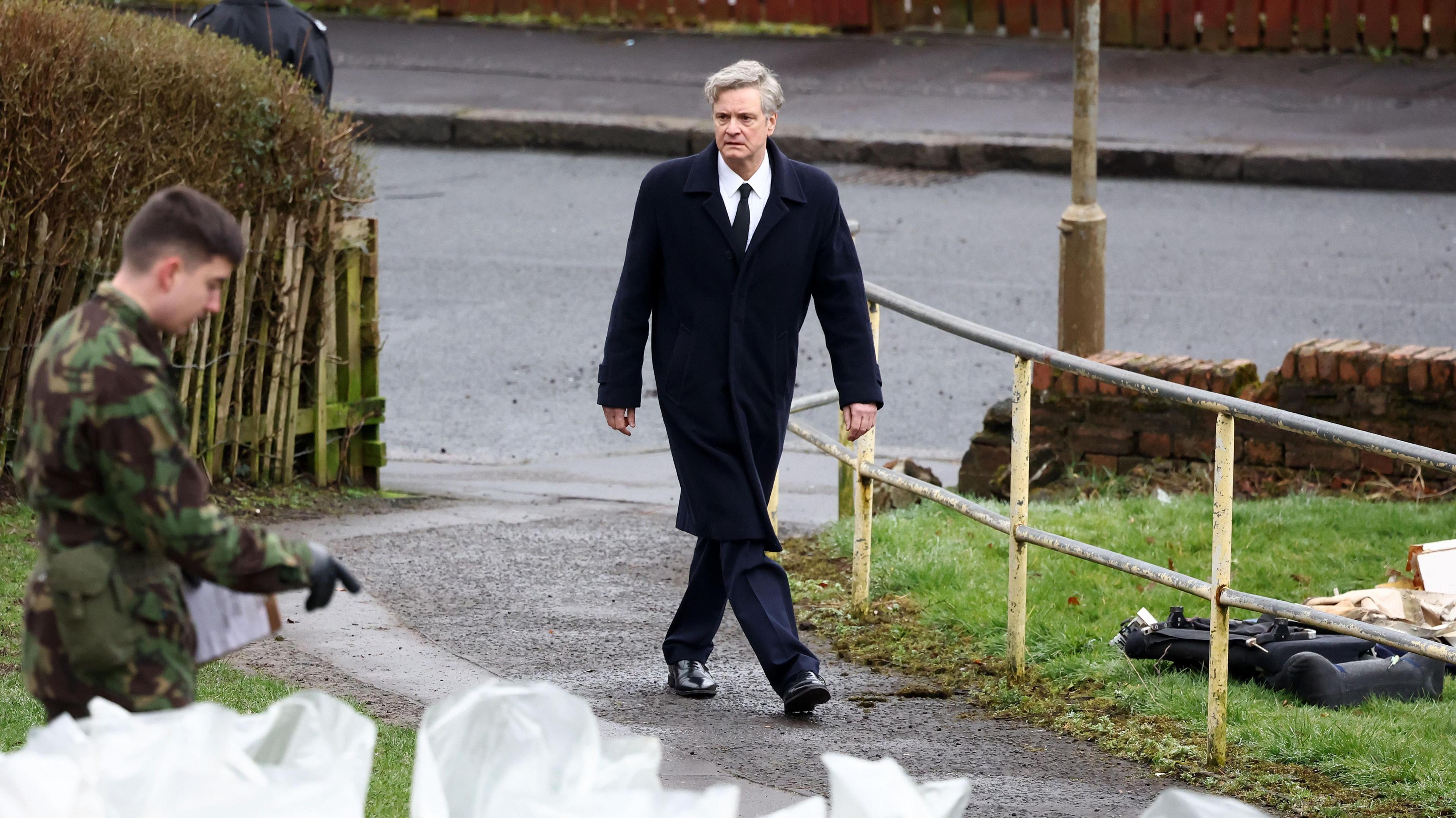 Colin Firth plays Dr Jim Swire in a new TV series. The photo shows Colin Firth in character, in a black suit and tie, walking down a path. There is a man in an army uniform to his left and pieces of white rubble from the plane are at the bottom-left of the image.