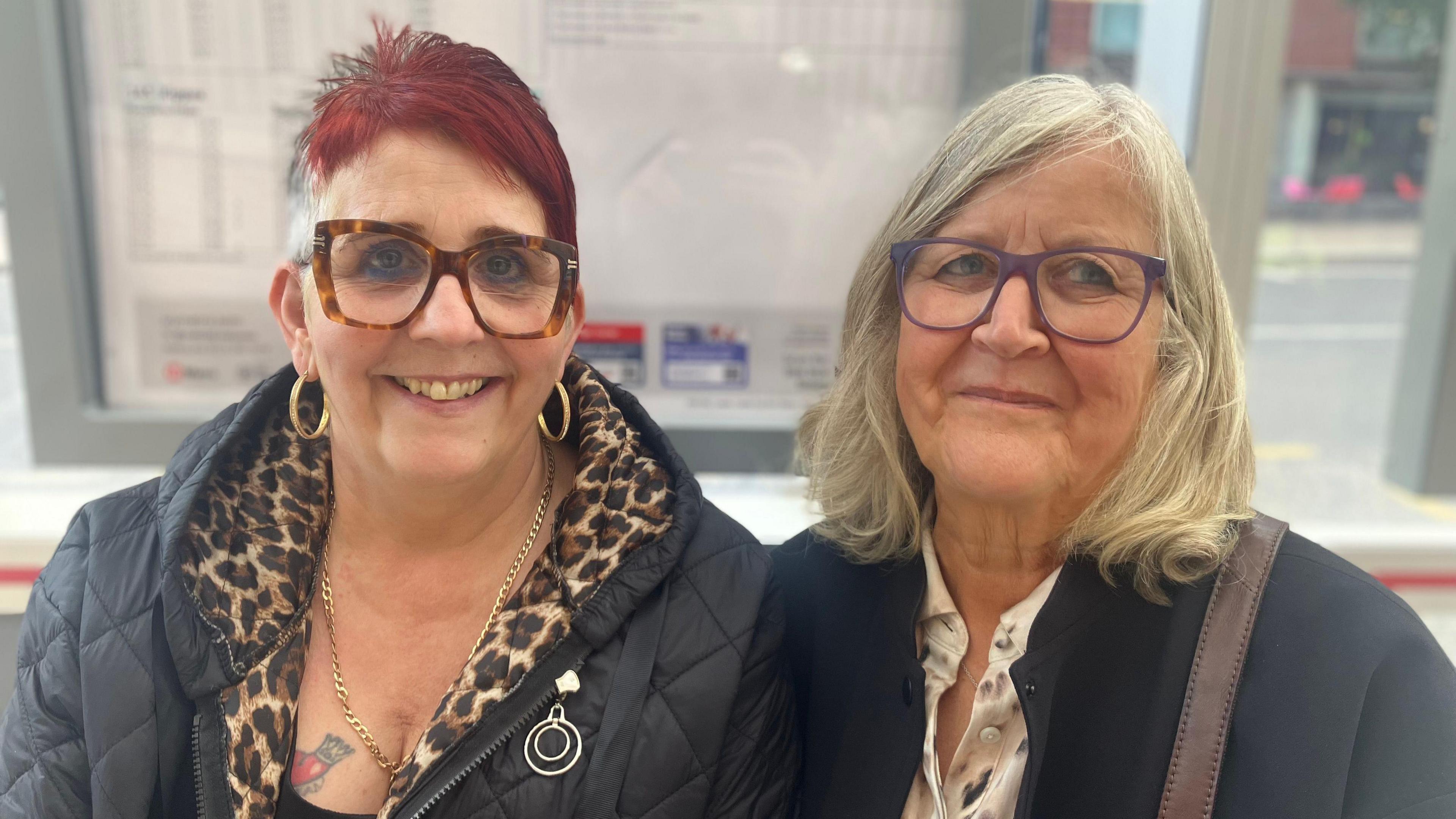 Two women sit at a bus stop in Leeds. The one on the left has short red hair, big brown glasses and gold hoop earrings. The one on the right has white blonde hair and wears a black coat with a brown bag over her shoulder.