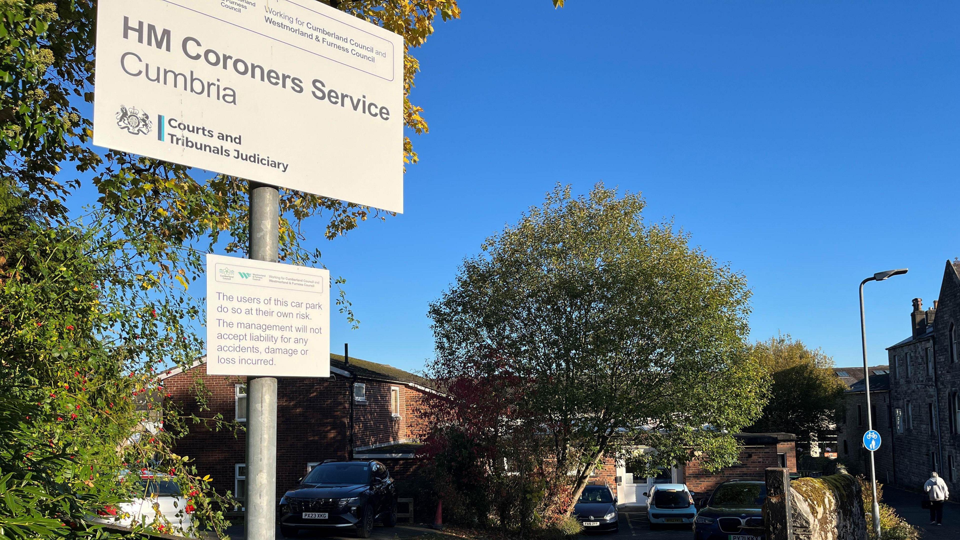 A general view of the access to the Coroners Court in Cockermouth. A sign reading "HM Coroners Service Cumbria" is in the foreground. Behind it is a small car park with a few cars parked in front of the court, which is a small red-brick building with a white PVC door.