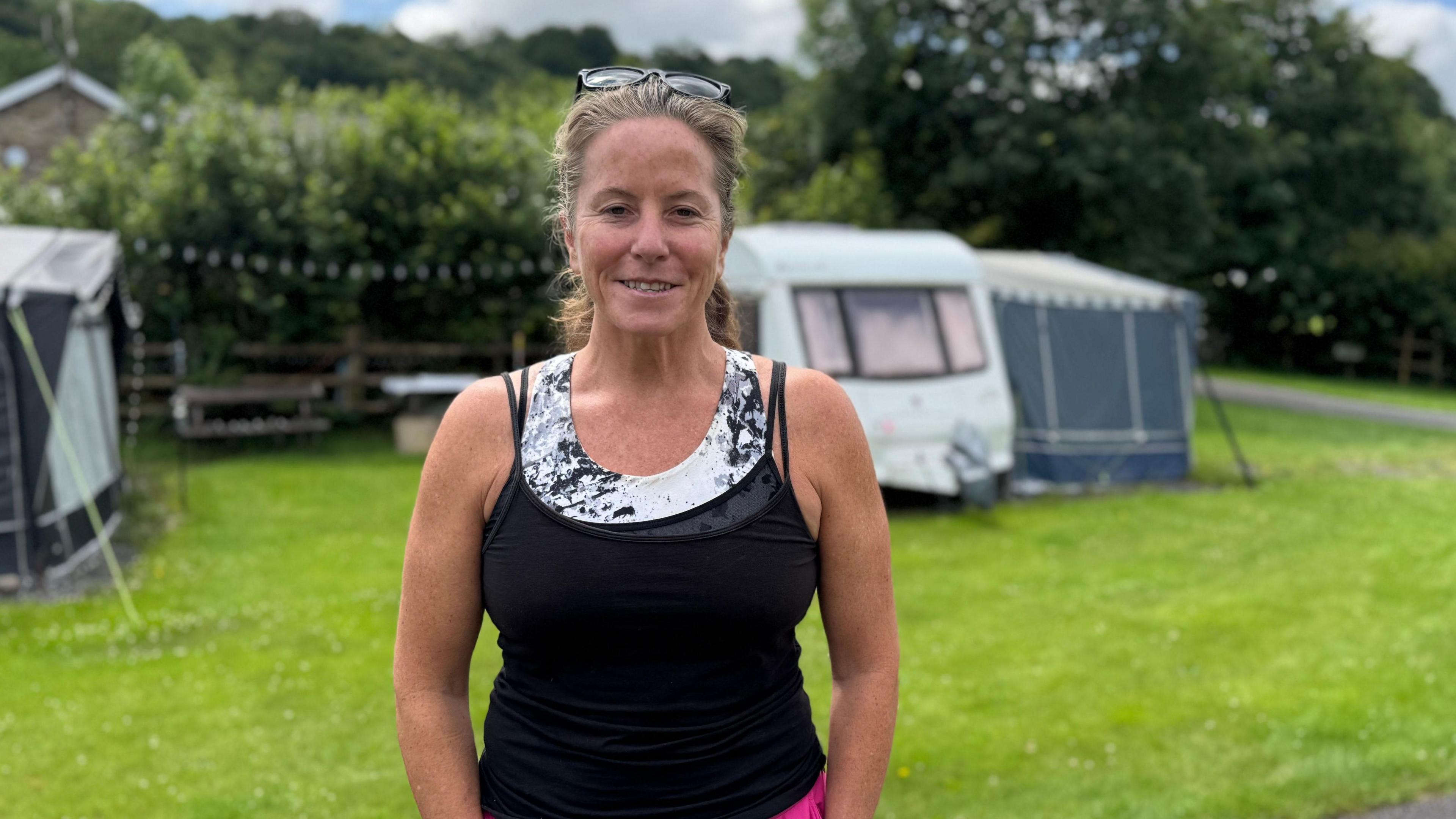 A woman wearing a black vest top and pink shorts. She has tied-back brown hair and sunglasses on her head. She is smiling and looking at the camera. Behind her is a caravan on a grassy field.