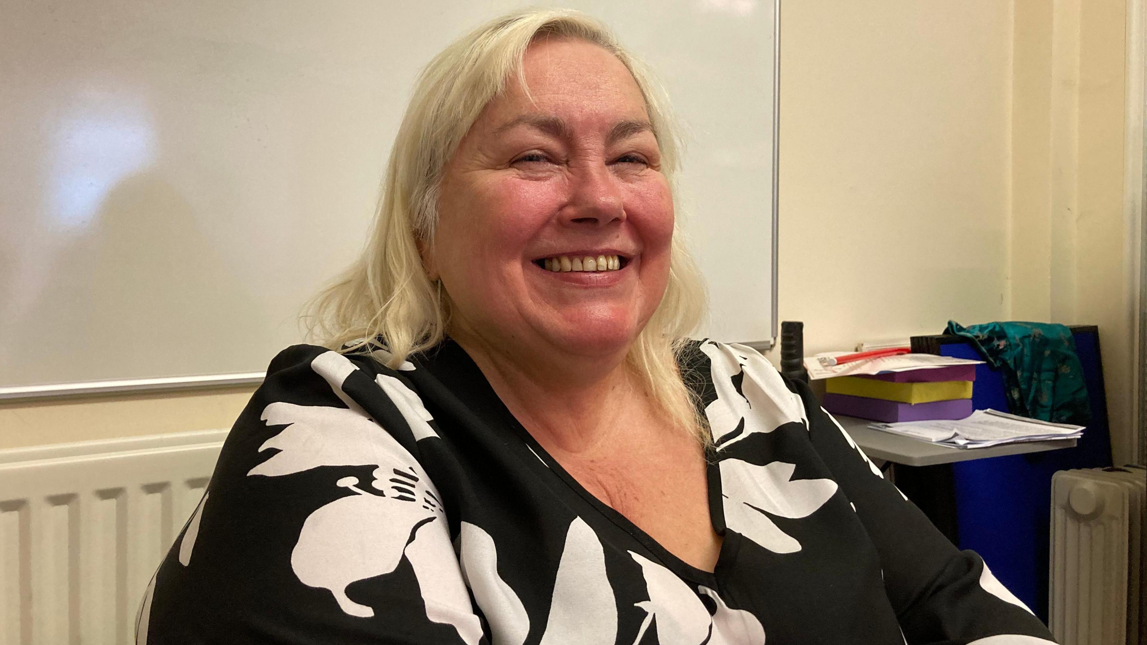 A woman with long blonde hair and a black and white top smiles as she sits in front of a whiteboard.