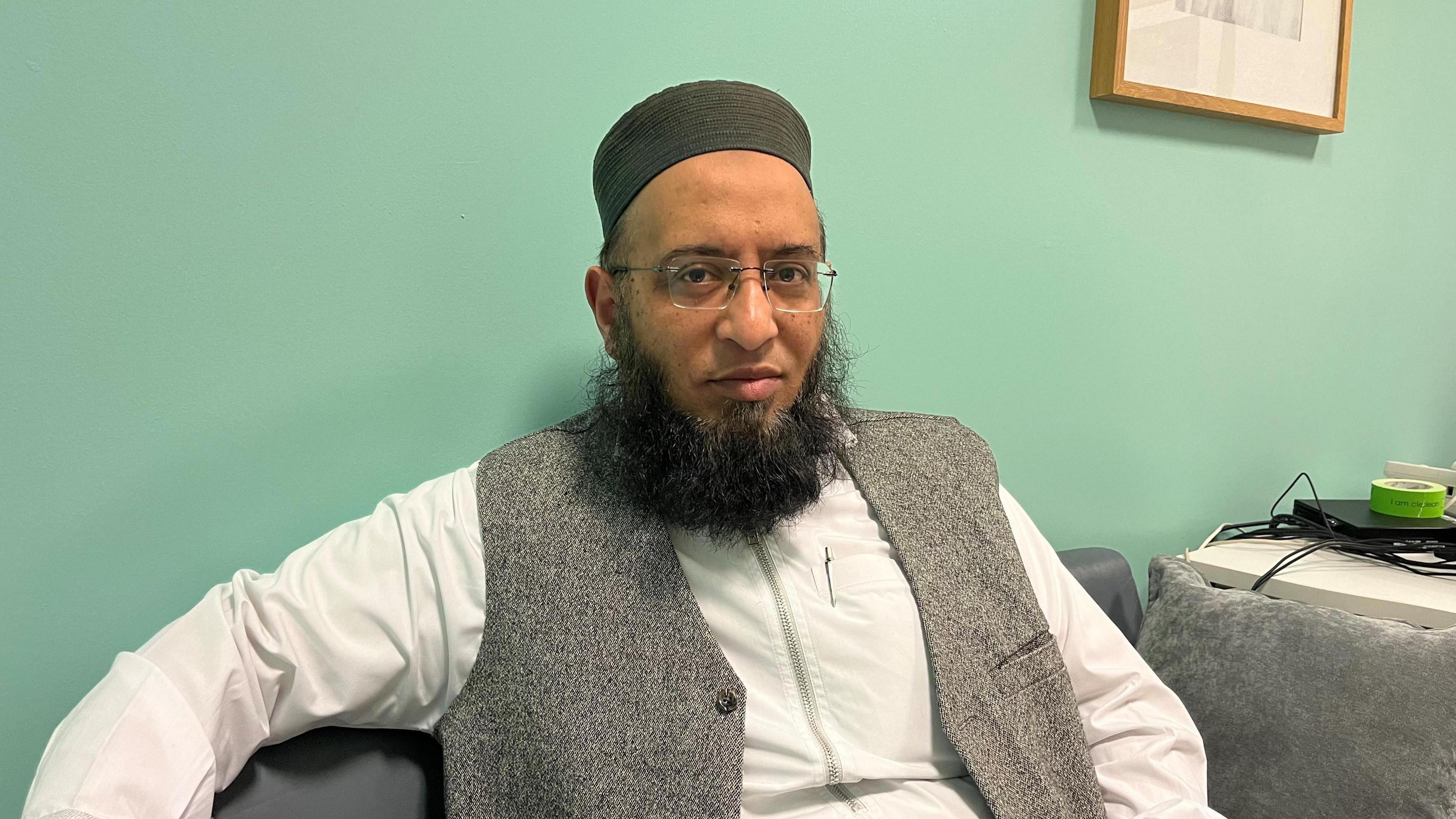 Mufti Zubair Butt wearing a white long sleeved shirt and grey waistcoat looks at the camera