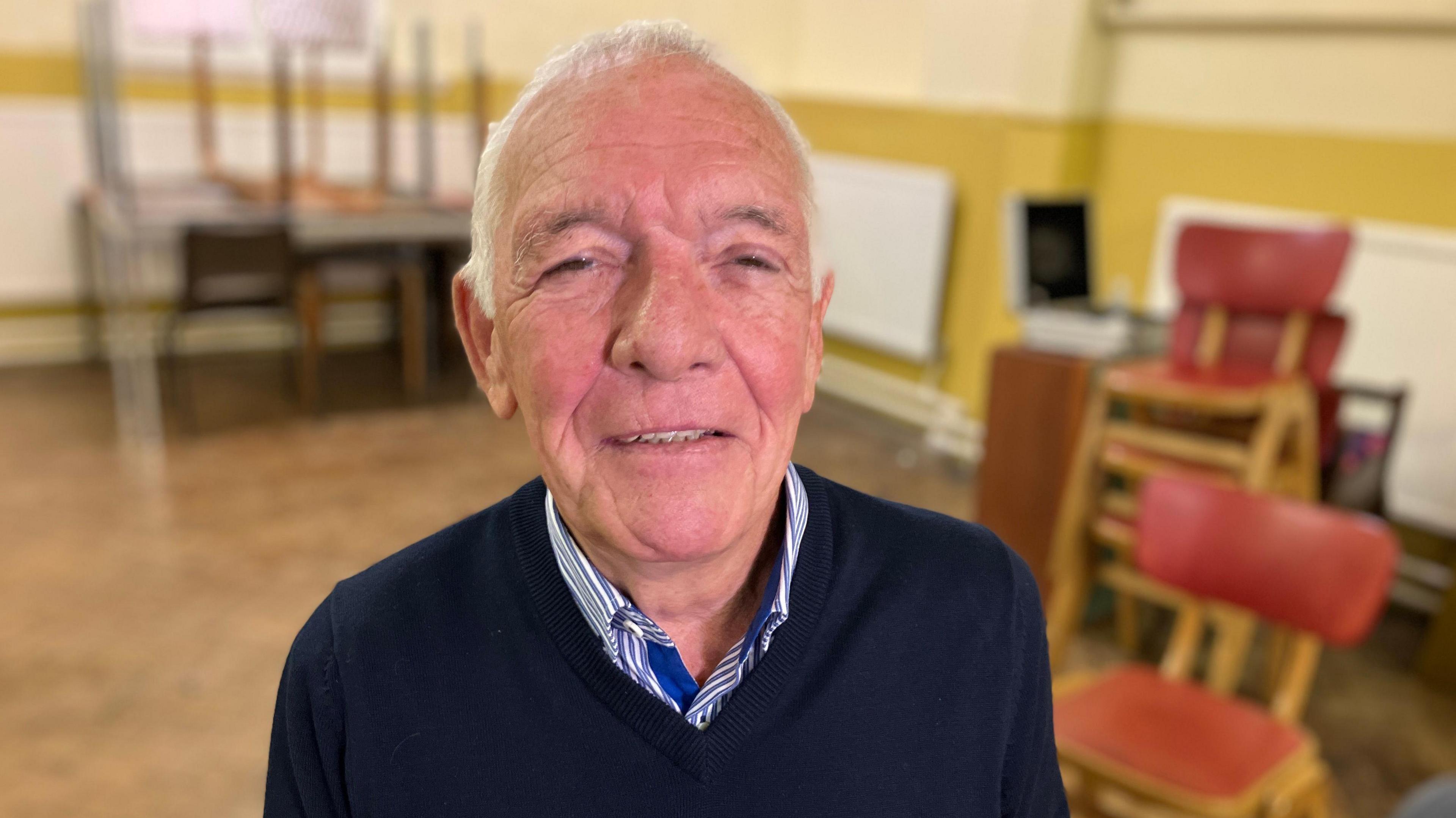 An older male  with thinning white hair is centre of the photo wearing a navy blue jumper and the white and blue striped collar of his shirt is also visible. Behind him and out of focus are mustard and cream colour walls, some wooden red chair are also stacked over his left shoulder and there are some tables stacked in the background over his right shoulder.