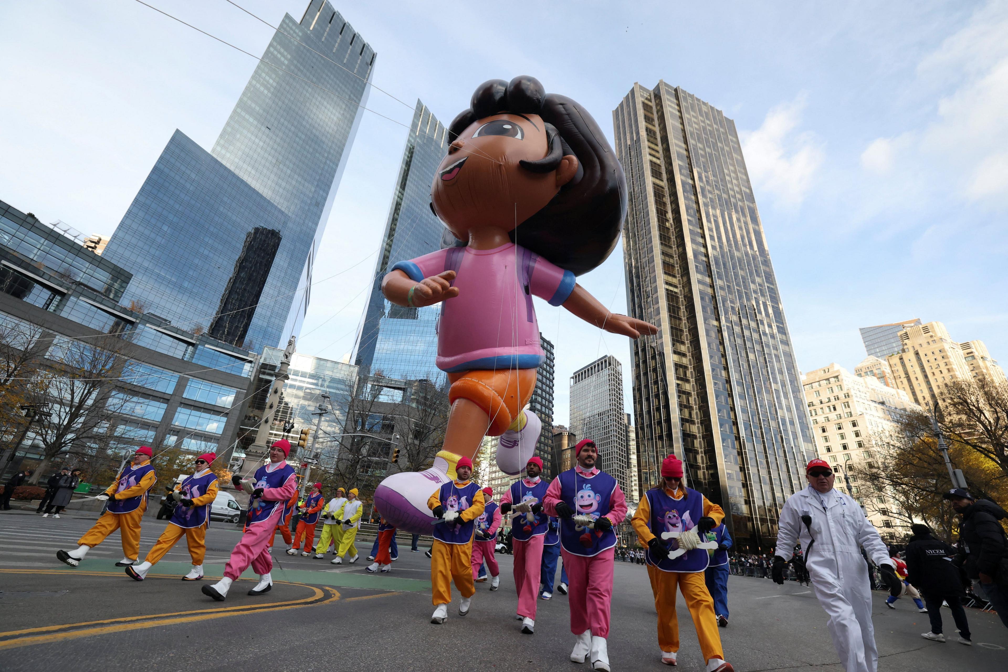A giant Dora the Explorer float is held by people wearing pink and orange, wearing purple tabards.