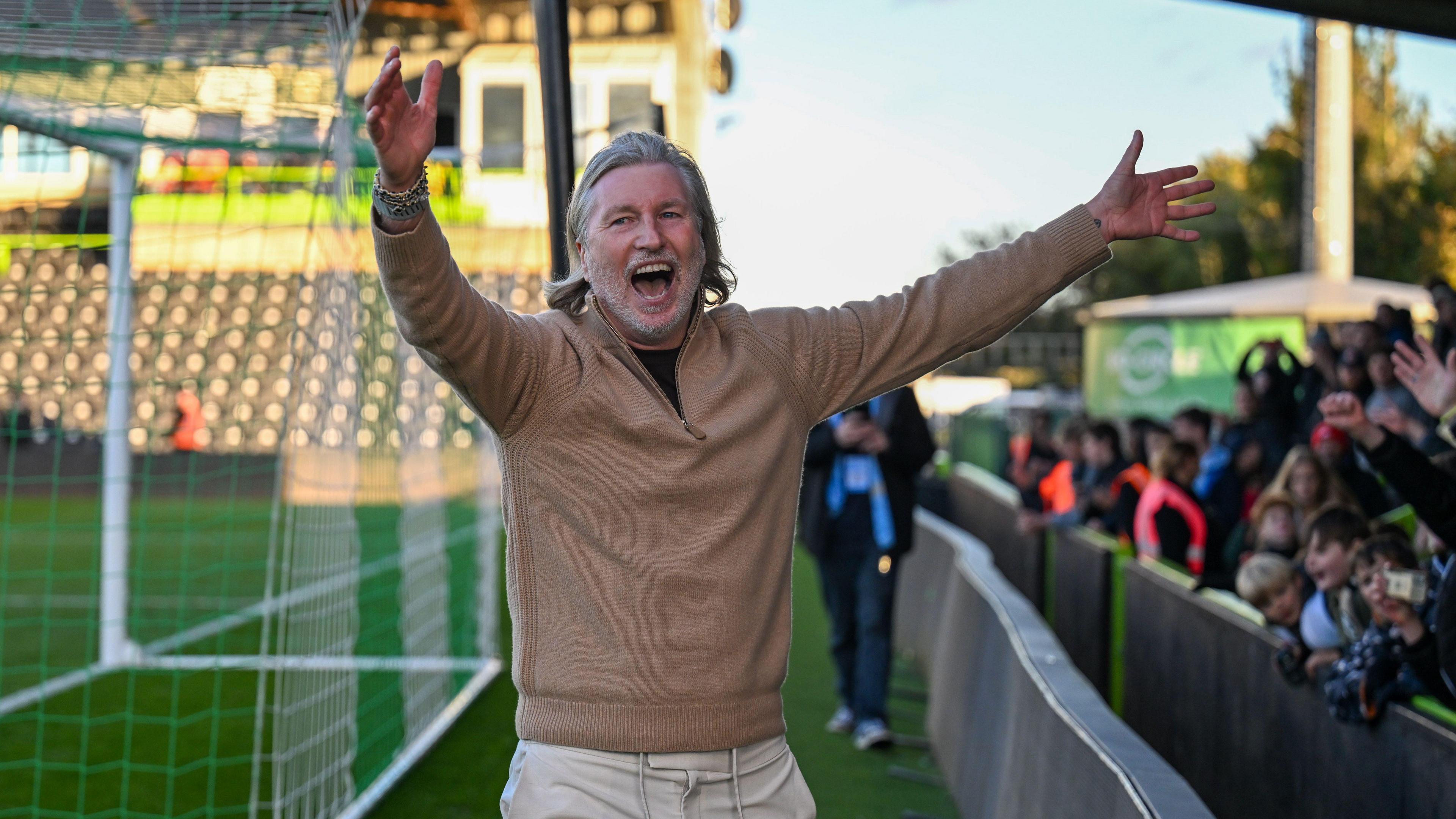Robbie Savage holds his arms in the air and smiles as he celebrates in front of fans on the edge of the pitch