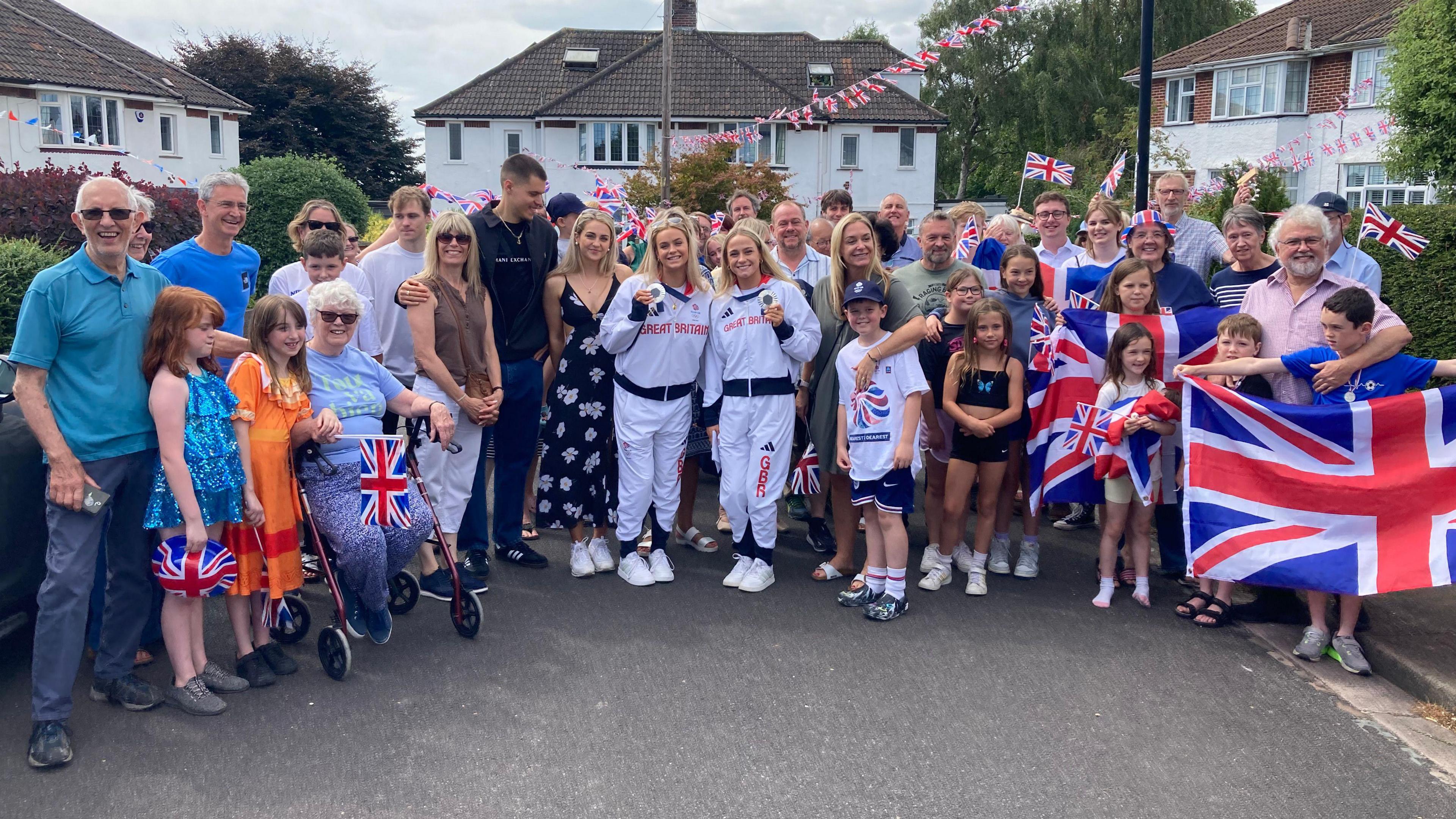 A wider shot of Kate and Isabelle wearing Team GB tracksuits wearing their silver medals, surronded by their family, friends and neighbours, outside on their cul-de-sac