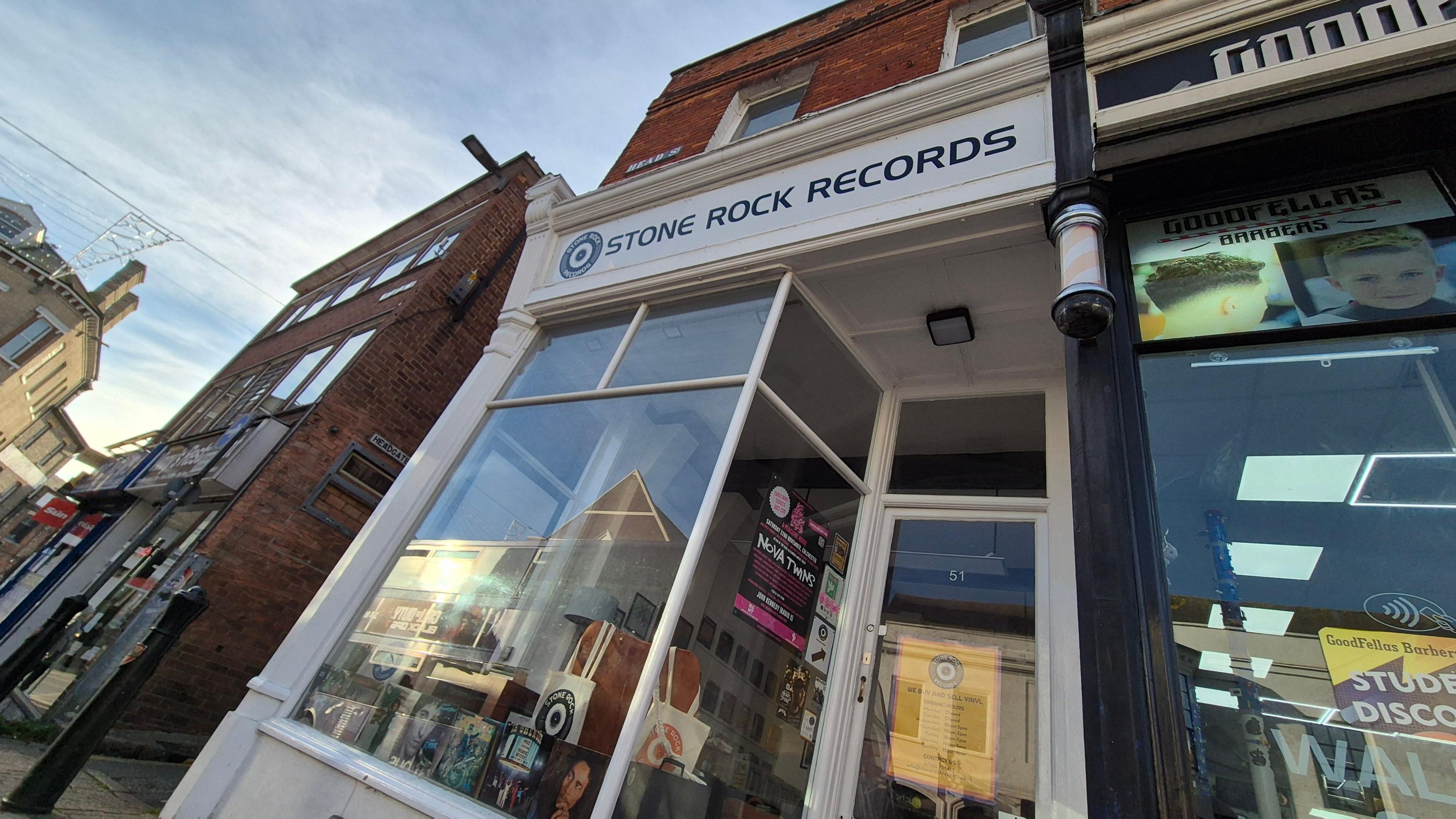 The outside of a record shop, with a white front, and posters and records in the window. Other shops are either side of it.