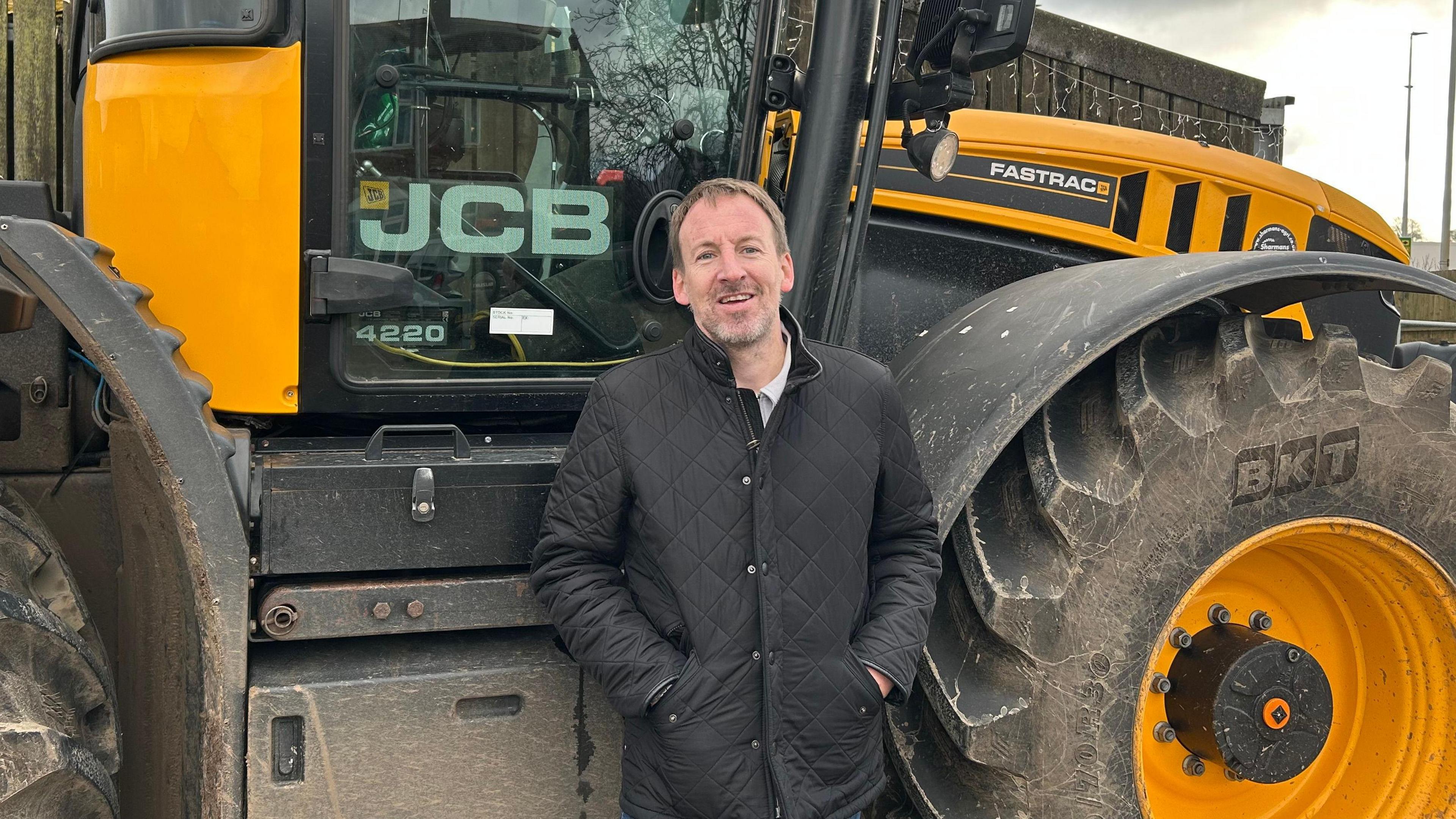 Ryan Archer stood in a black jacket in front of a yellow JCB tractor.