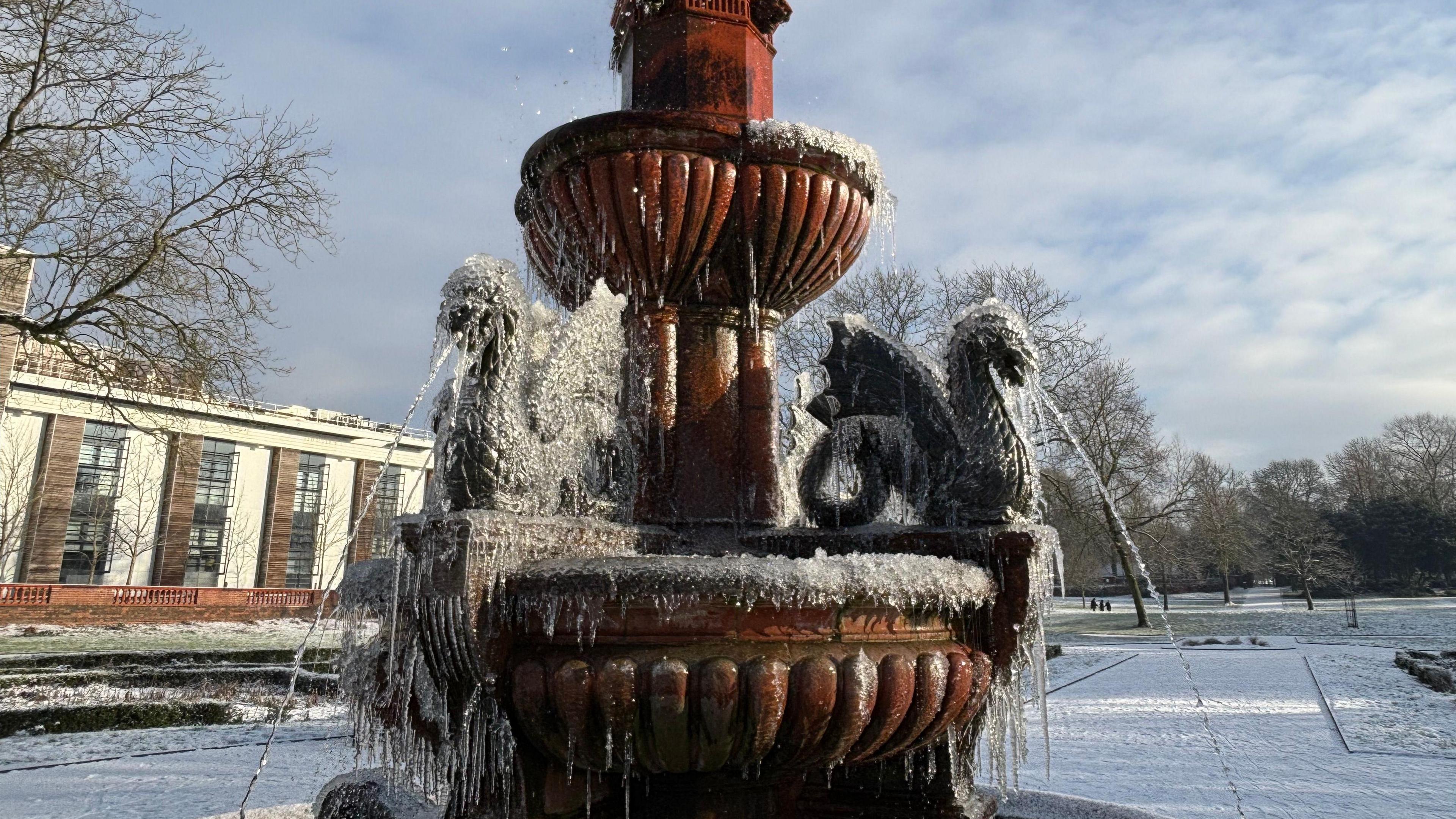 A fountain with statues of dragons on spouting water. The water has frozen and icicles are hanging off the dragons and the fountain. A park is visible in the background as is a building to the left.