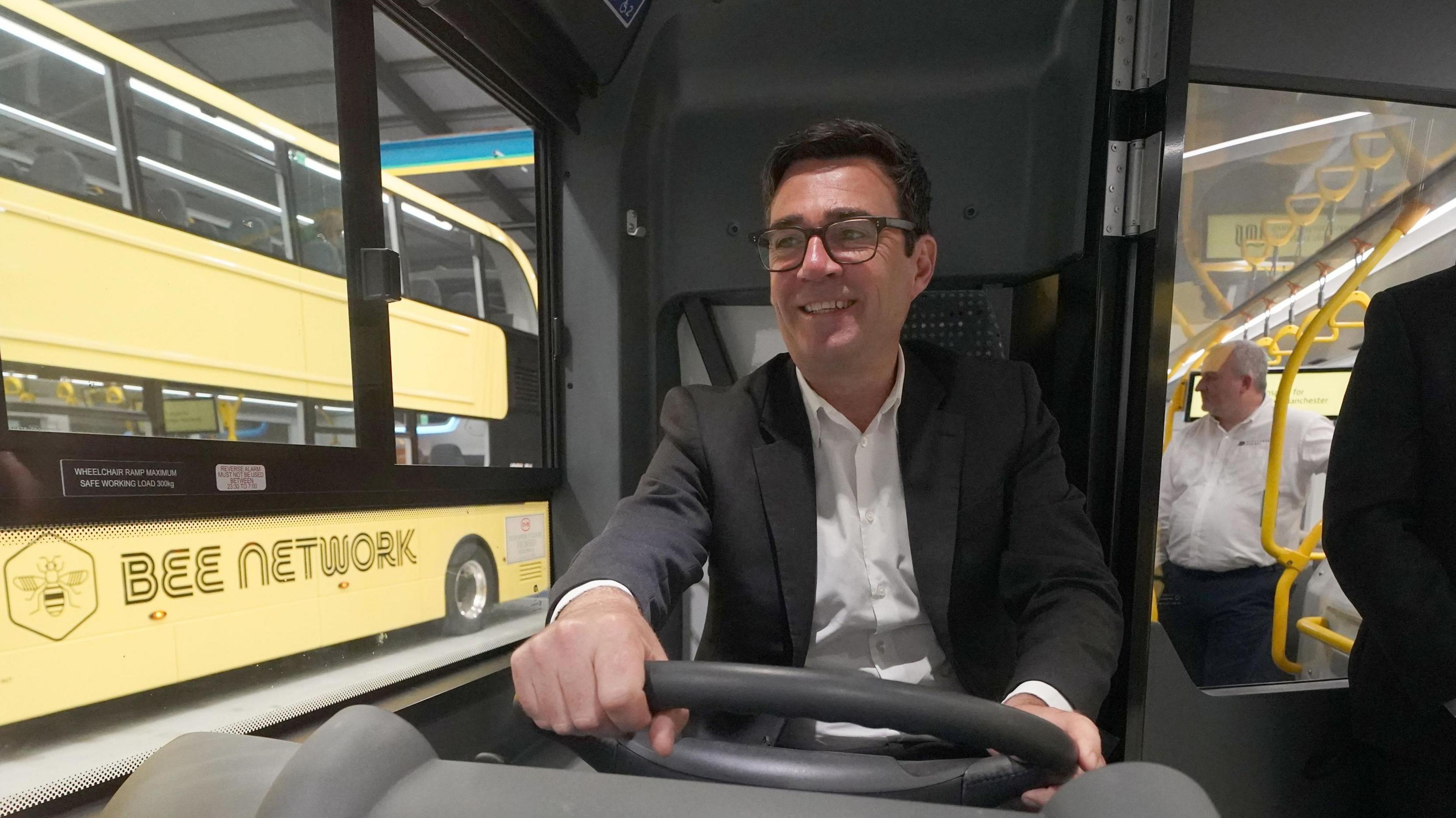 Greater Manchester mayor Andy Burnham smiles as he drives a Bee bus - a yellow double decker bee bus is out of the window