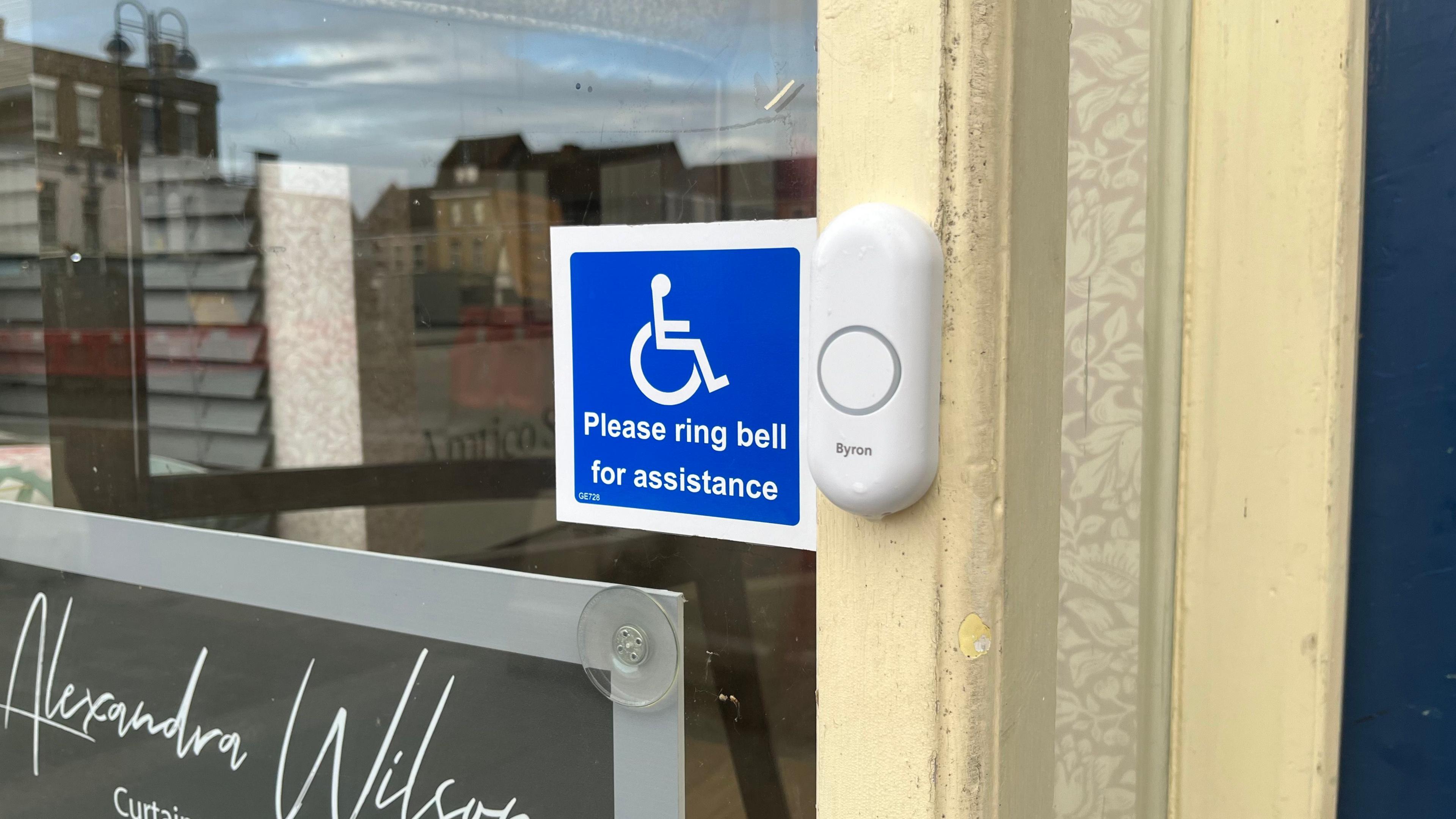 A white doorbell on a yellow painted doorway. On a window next to the bell is a blue and white sticker with an stick image of a person in a wheelchair which reads 'please ring bell for assistance'.