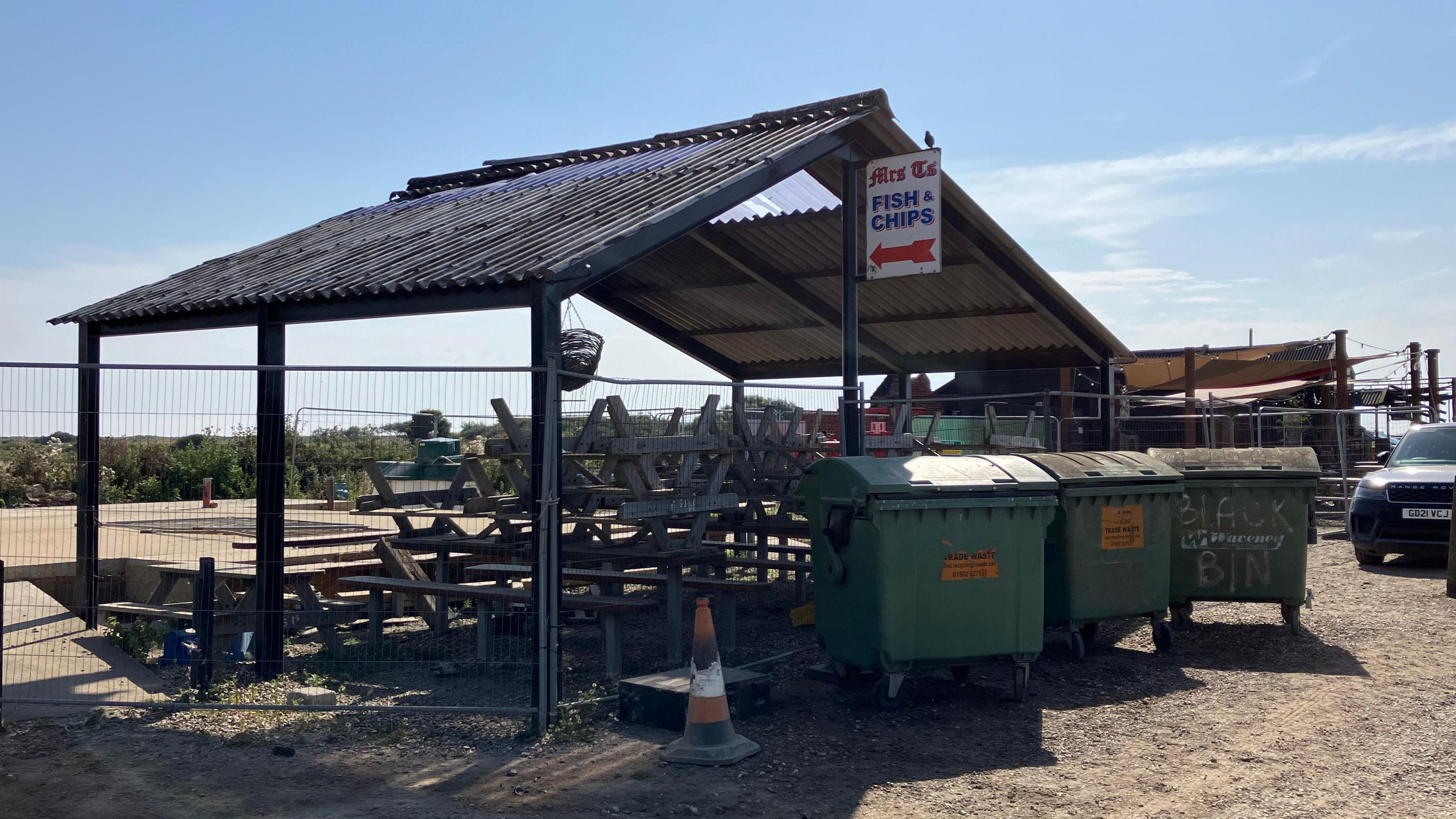 Stacked up seating benches and green wheelie bins can be seen at the area which was destroyed by fire last May