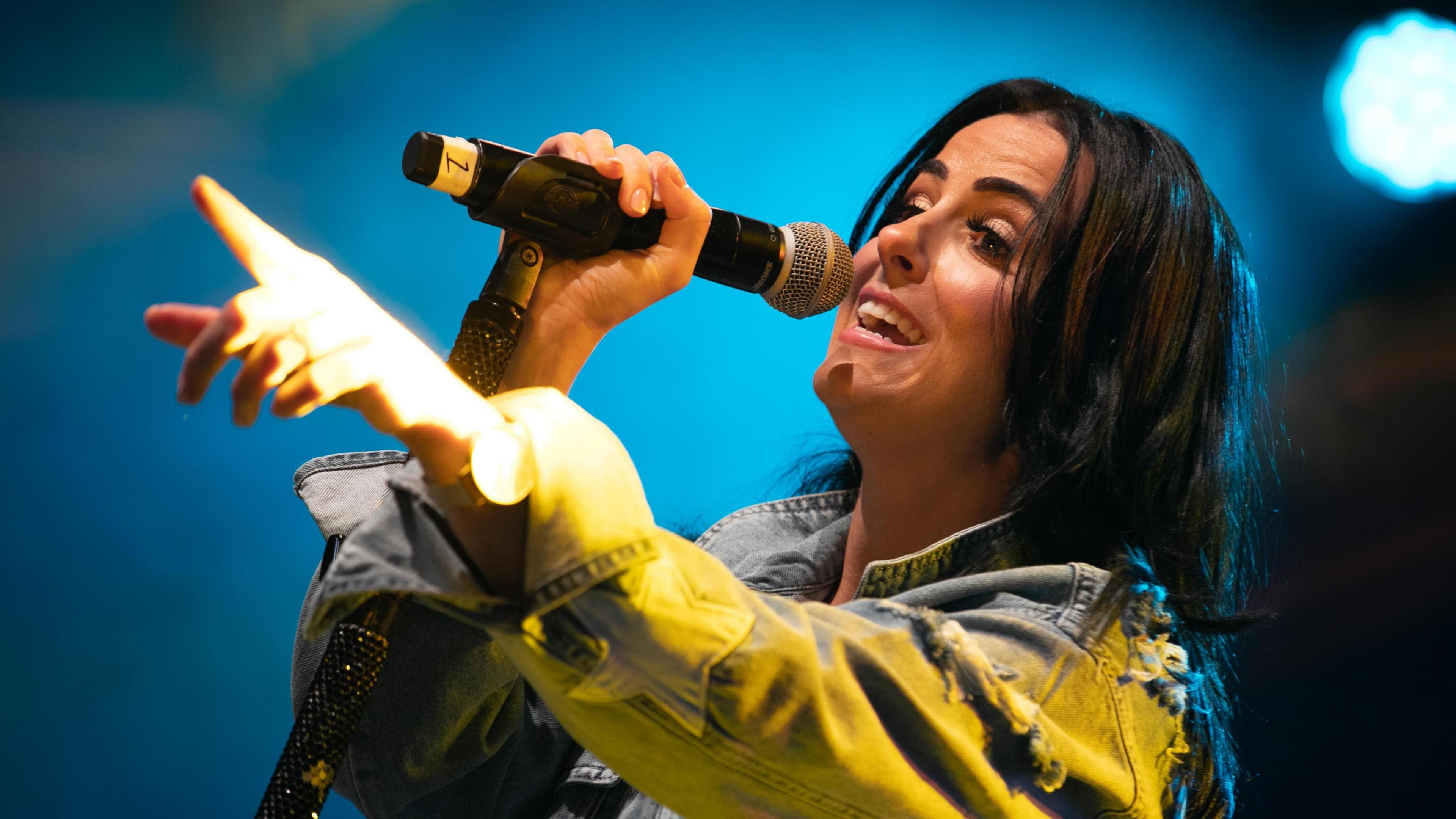 A woman with long brown hair singing into a mic as she wears a blue denim jacket.