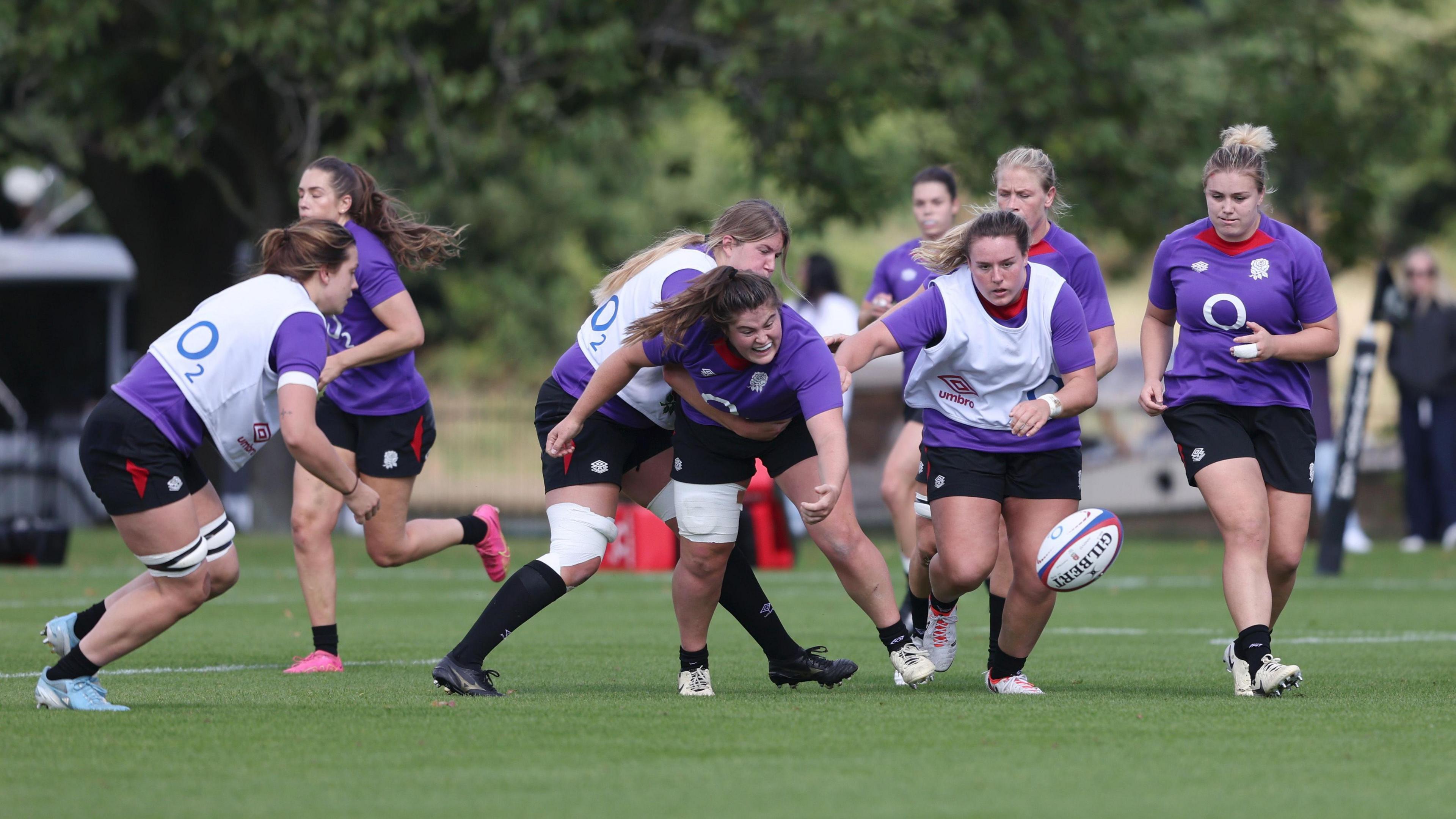 Maud Muir in the heat of a rugby game. Several players are running to the oval-shaped ball. Another player is holding Muir across the waist. Muir is seen wearing a mouthguard. It is a cloudy day.