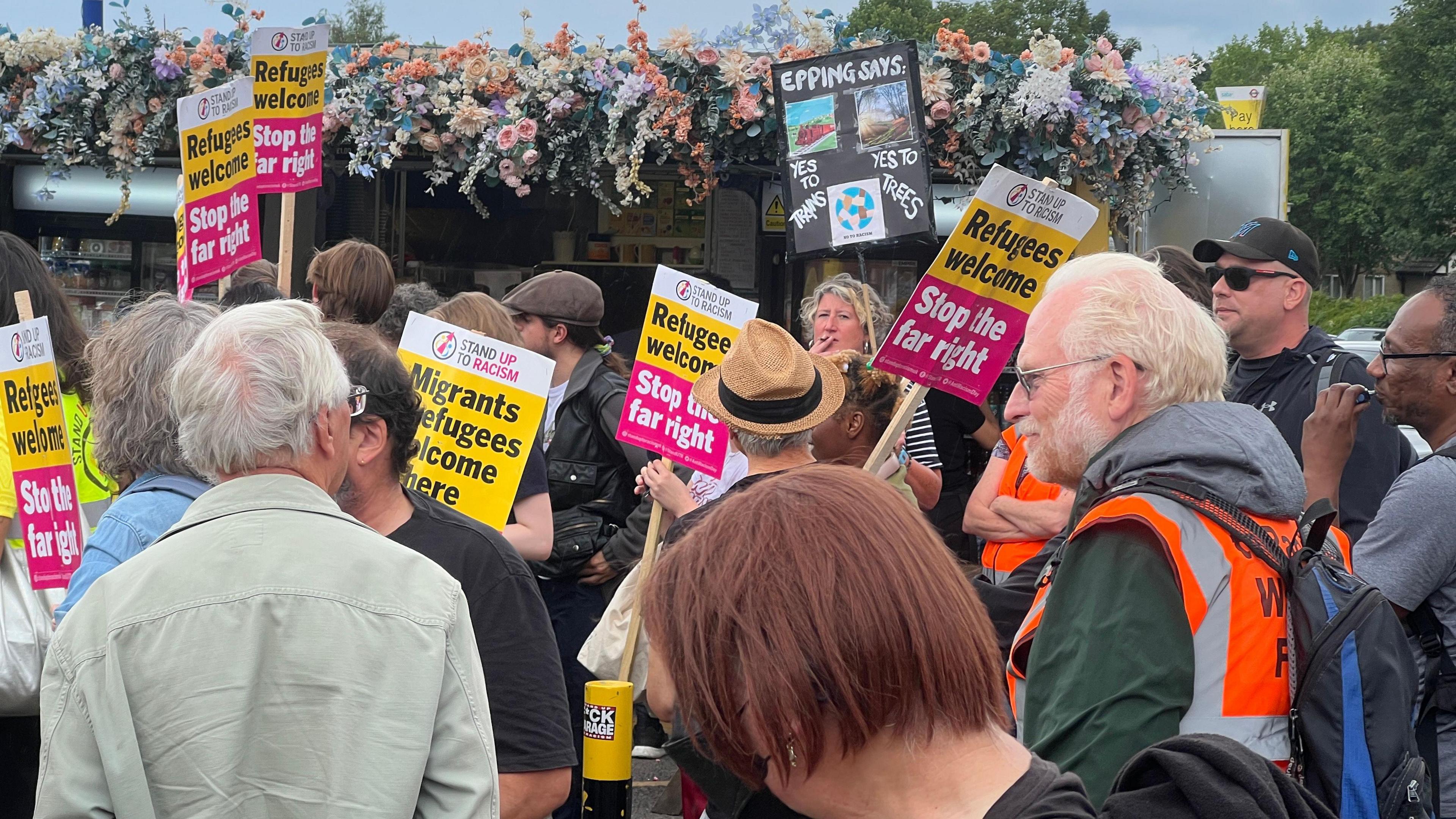 A group of people stand together with some holding banners that read "Refugees Welcome - Stop the far right". They are standing beside a building with flowers on the roof, and a green space behind it.
