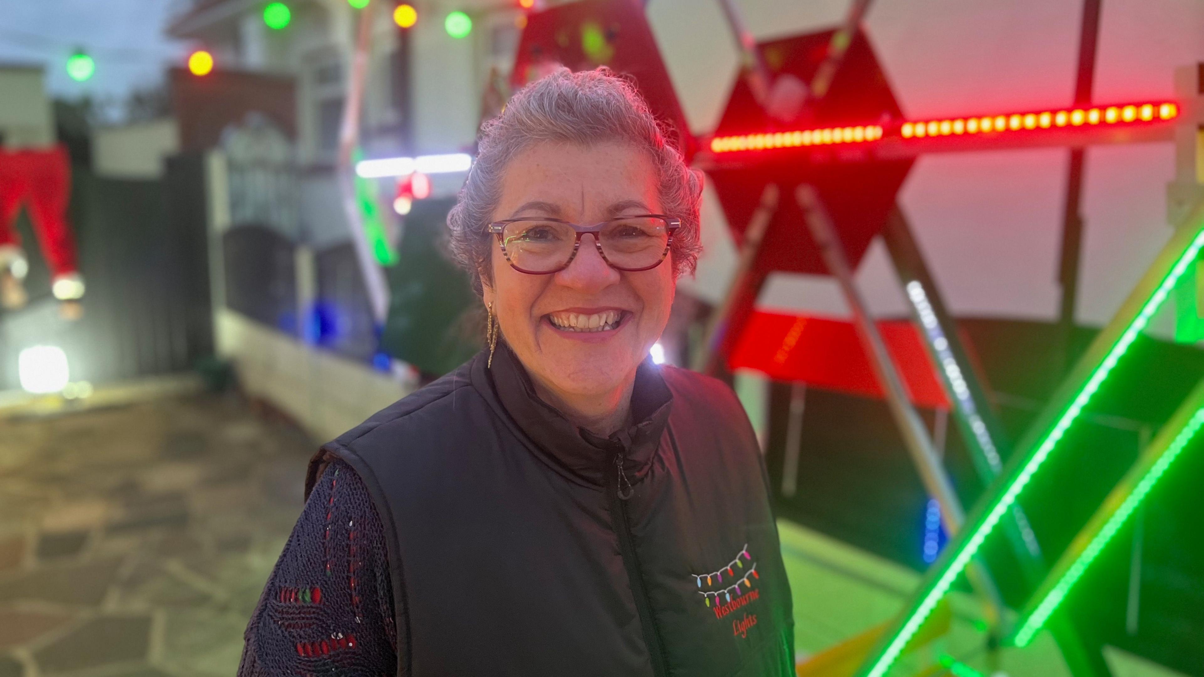 A woman smiling straight at the camera. She is standing in front of a Ferris wheel and is wearing a black gilet with the words "Westbourne Lights" embroidered on the front.