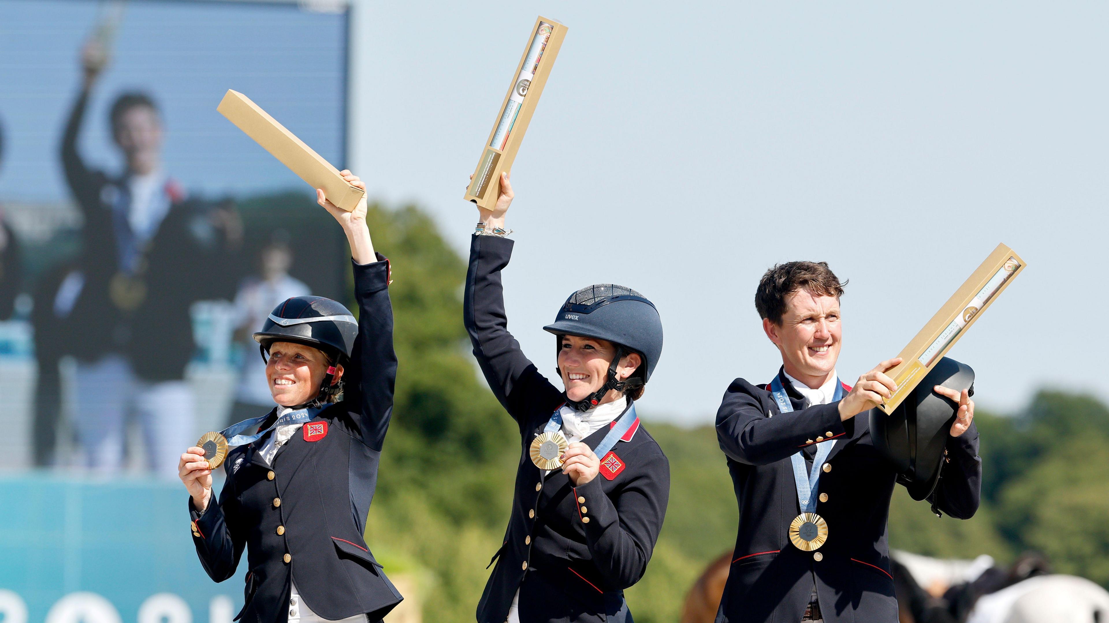 From left: Ros Canter, Laura Collett and Tom McEwen, wearing black equestrian jackets and riding hats, raise their Olympic diplomas to the crowd after being presented with their gold medals.