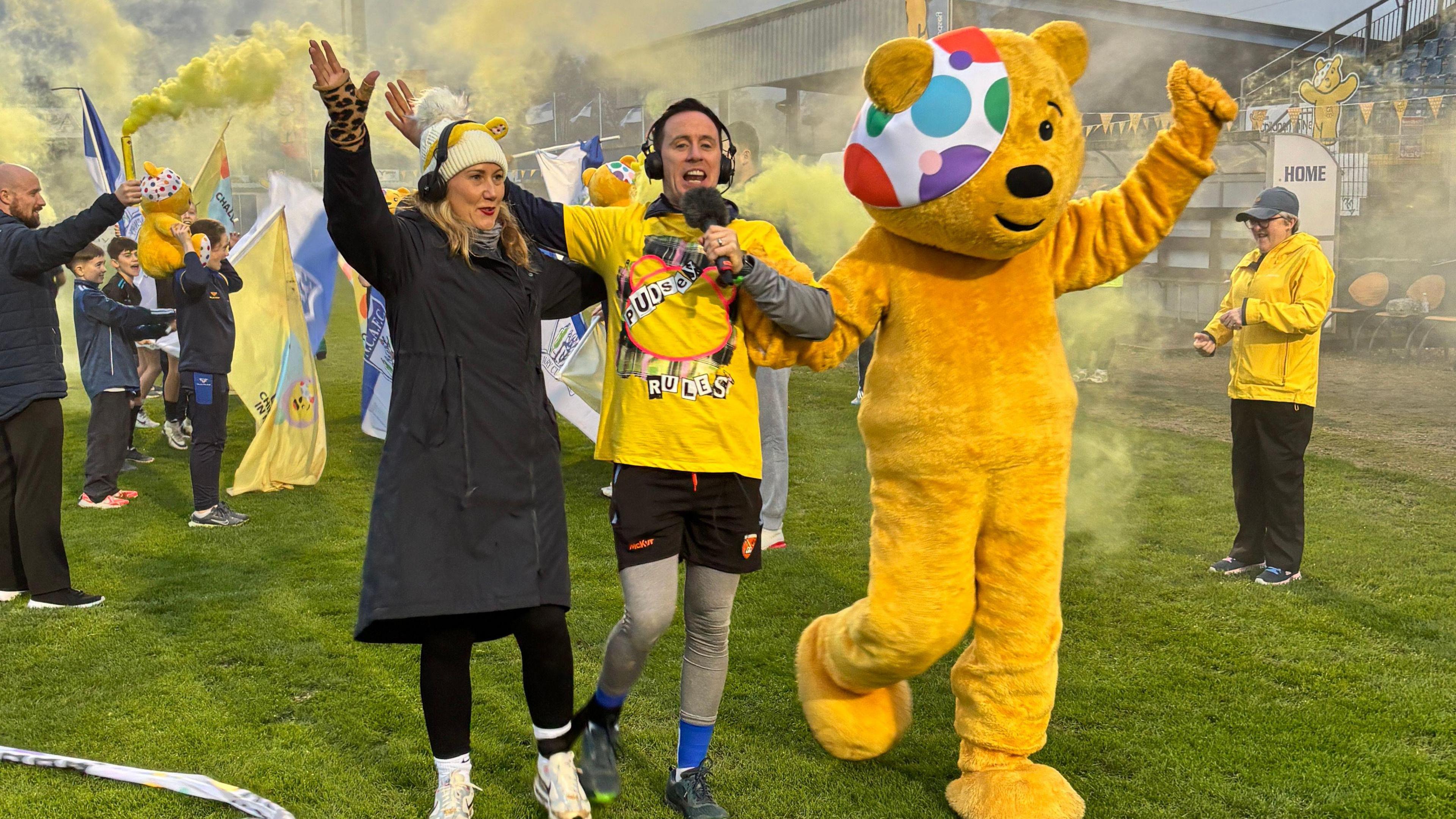 A woman and a man with a leg each tied together cross a finish line at the same time as Pudsey Bear in what looks like a sports stadium with crowds behind them - there are yellow flares everywhere in the background