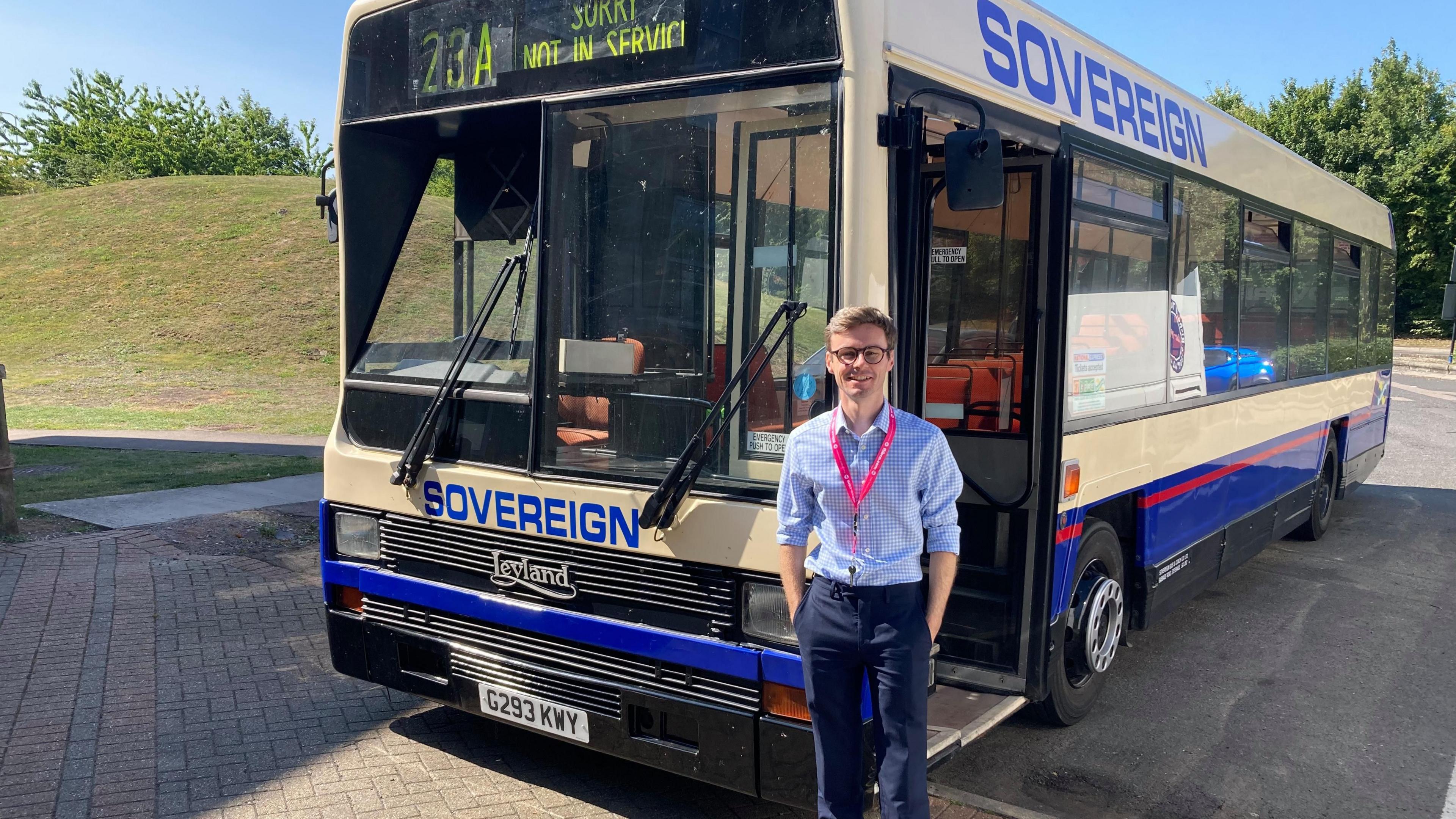 A man with short brown hair and glasses is stood in front of a white and blue bus with the words "SOVEREIGN" printed on in blue font.