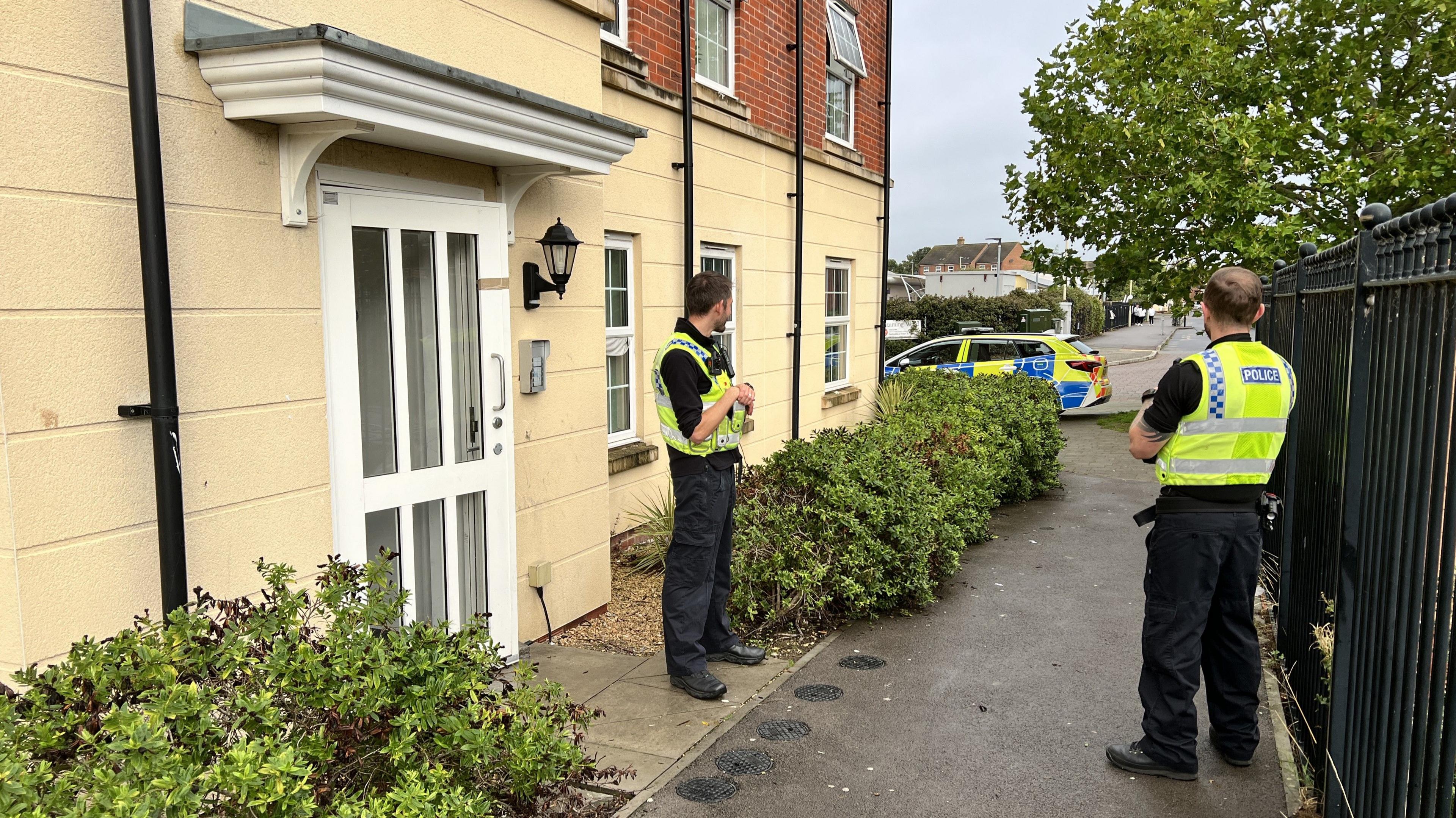 Two police officers wearing hi-vis yellow vests and black trousers and boots, standing outside the front door of a residential block of flats. There are bushes planted outside the windows and a metal fence. In the distance you can see a police car parked on the road.