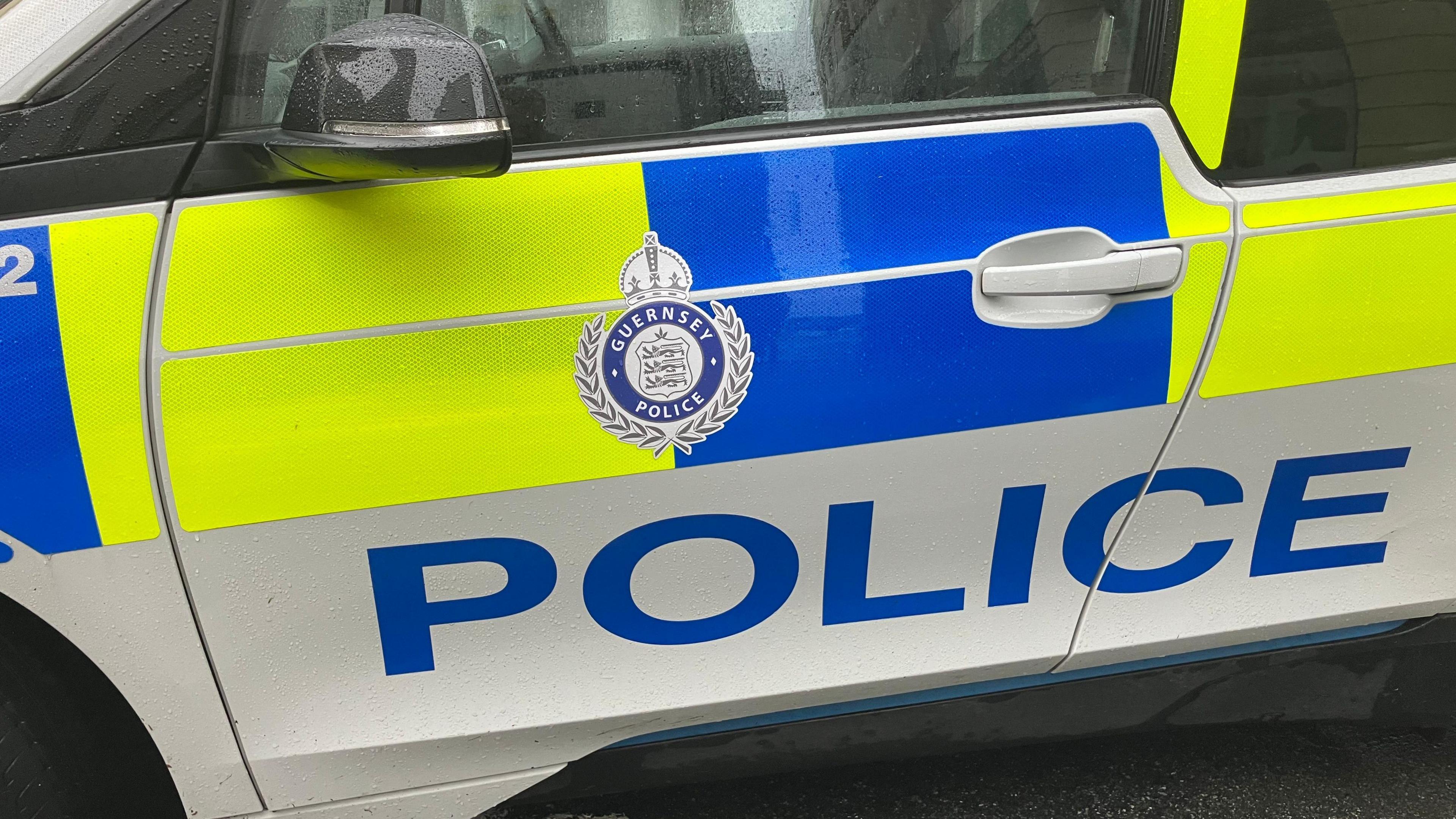 A close up shot of the door of a Guernsey Police car parked on a street. It is blue, white and yellow. It has police written in large blue block capitals on its side. Rain drops which have settled on the wing mirror, window and door are visible.