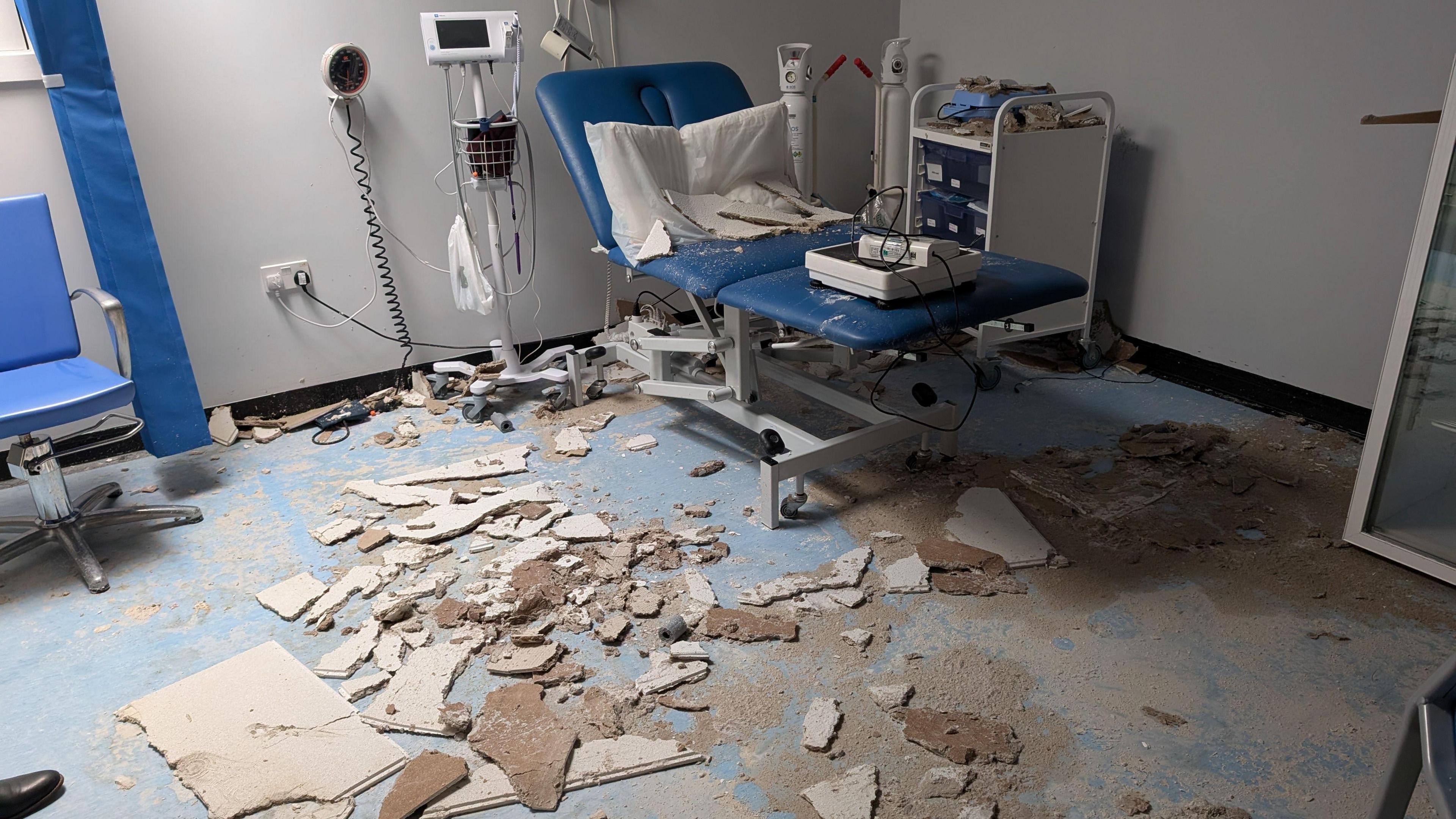 A view of the damaged room in the surgery. The roof has collapsed and parts of it is on the floor. There is a blue bed on the right as well as other equipment. 
