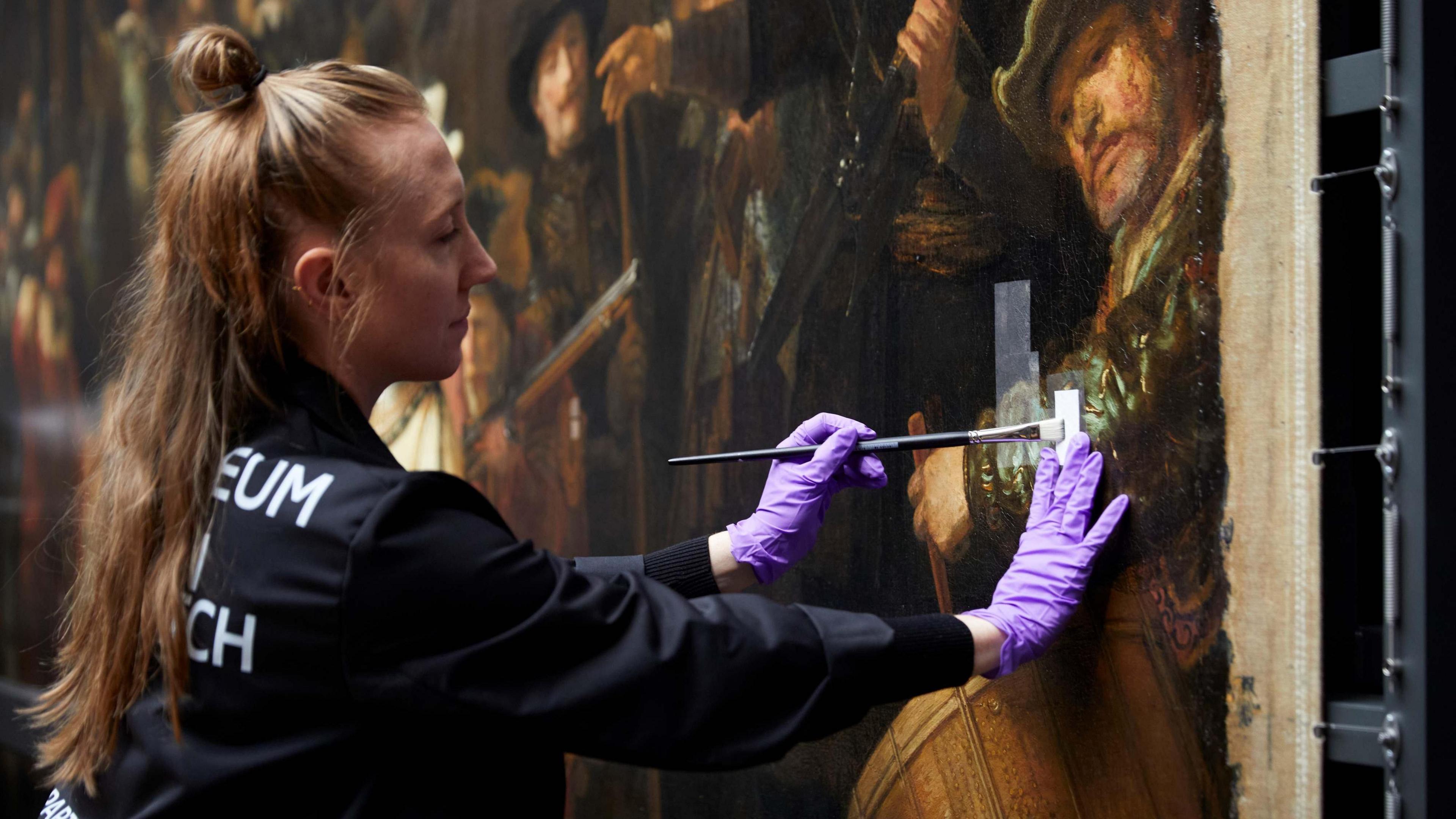 A woman with long blonde hair in a ponytail is wearing black protective clothing and purple latex gloves as she carefully removes the varnish from a section of The Night Watch