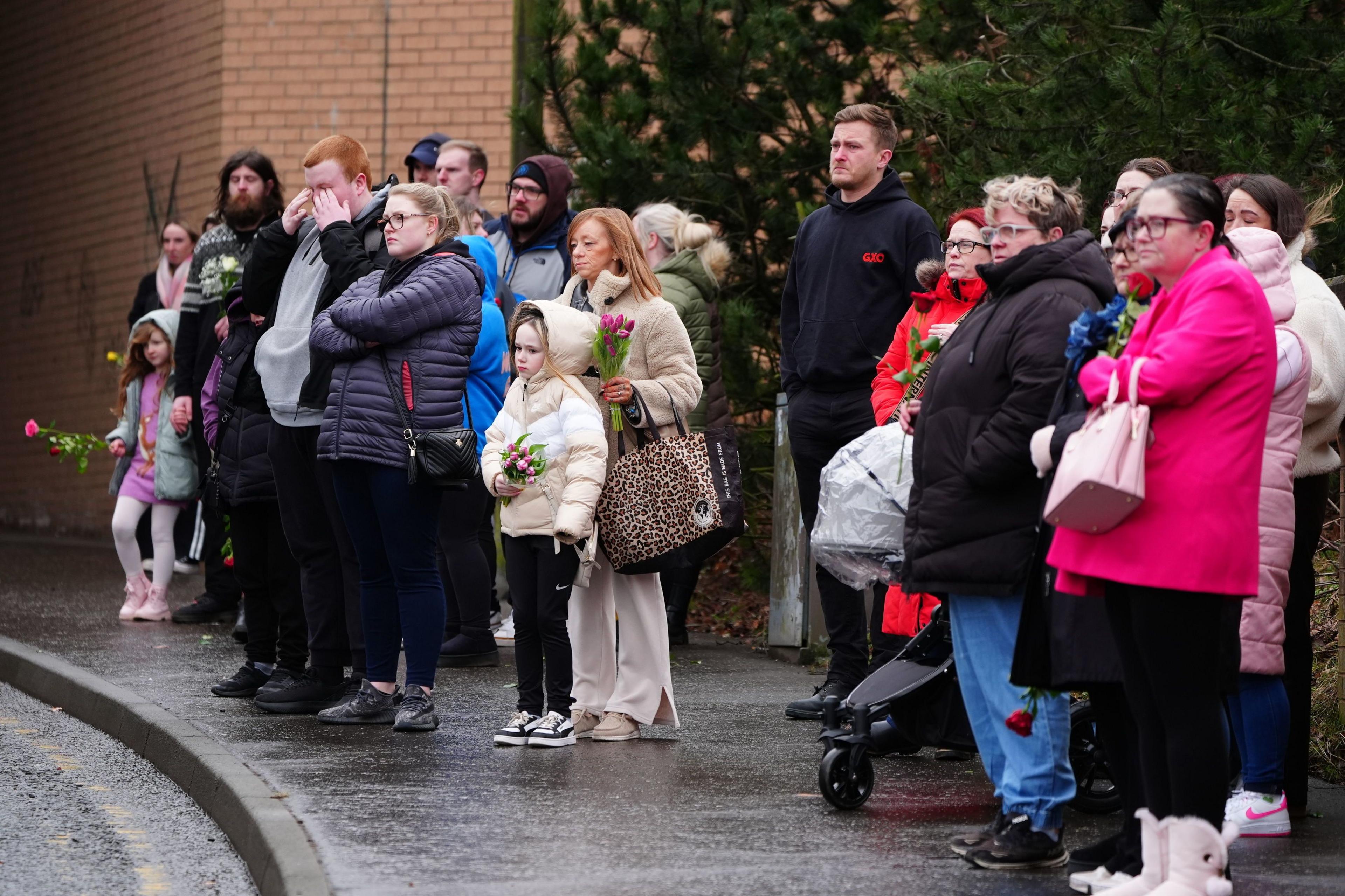 Several adults and children, some holding flowers, stand on a pavement watching a funeral procession. Many are wearing boots and heavy winter jackets.