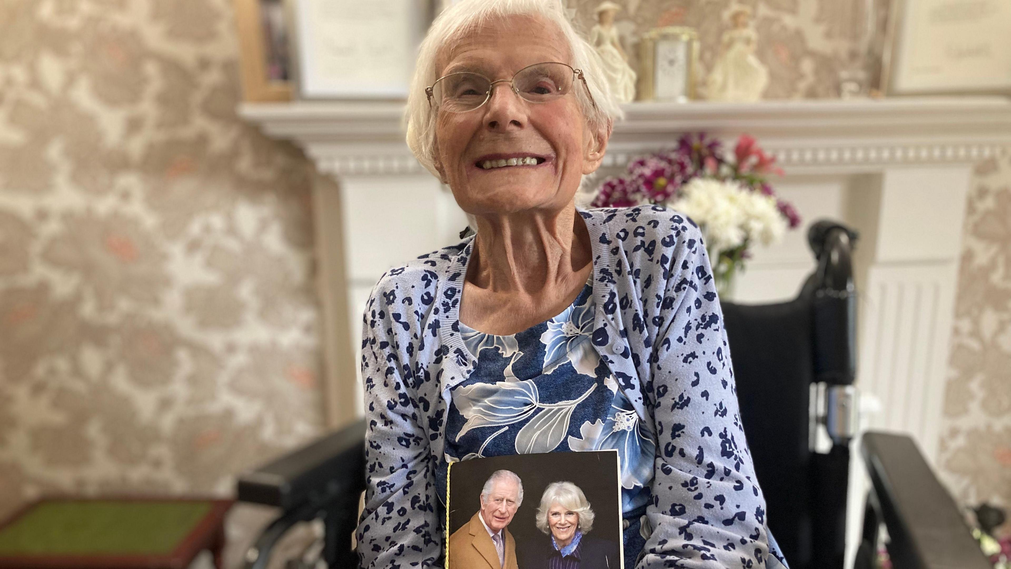 Elderly lady in a blue floral dress holding a card showing King Charles III and Queen Camilla. 