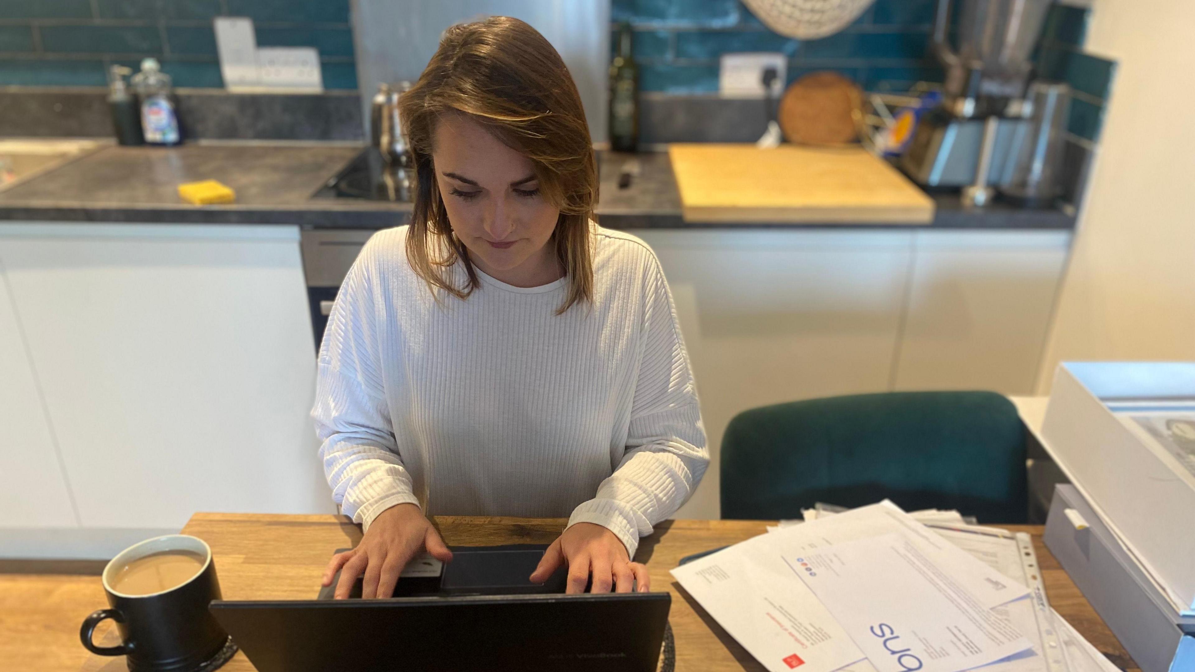 Lucy Tissington in a white top, sitting at a kitchen table on her laptop with a mug of tea and documents next to her