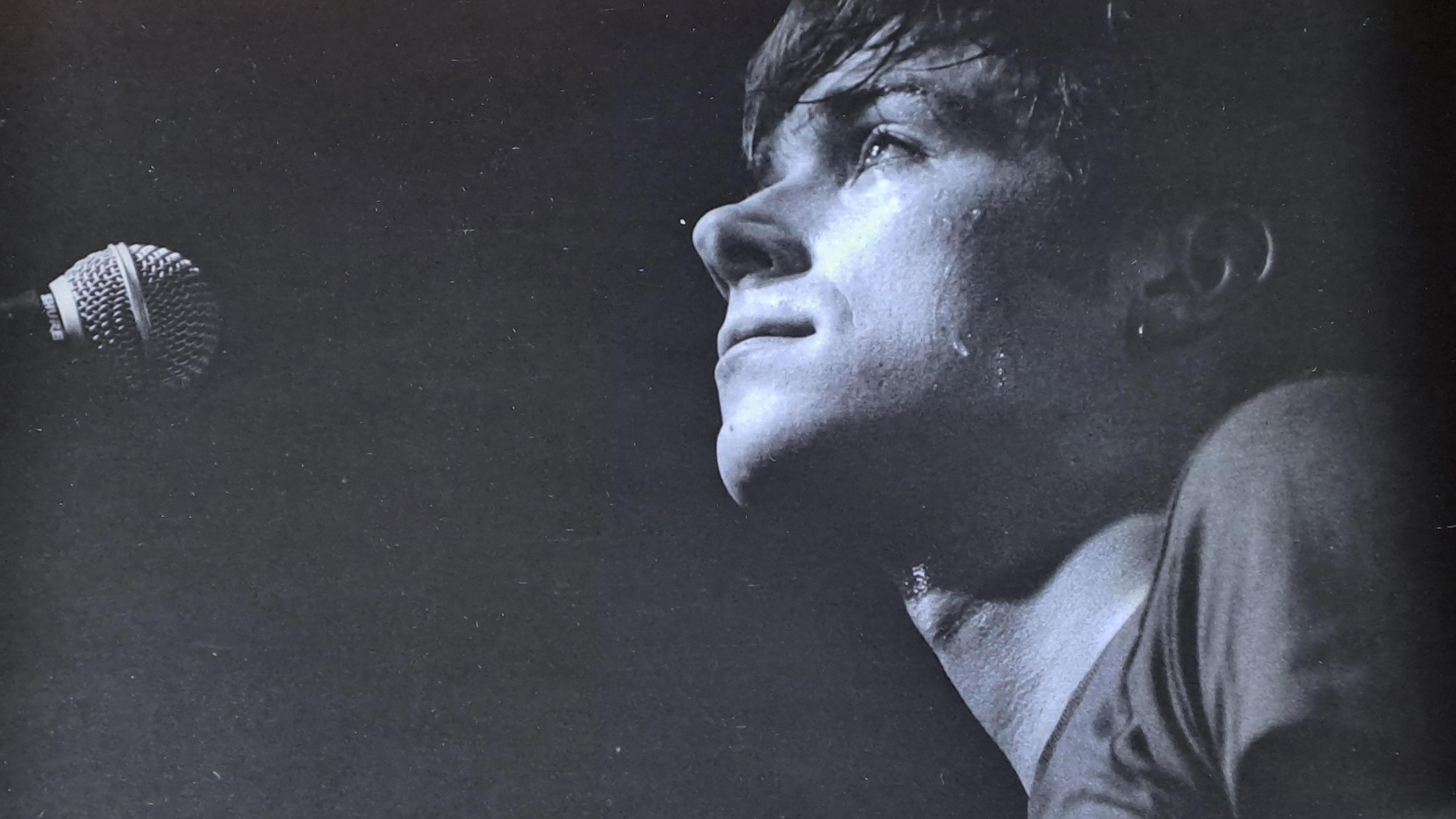A close-up image of young Damon Albarn who is looking up towards a ceiling and standing back from a microphone.