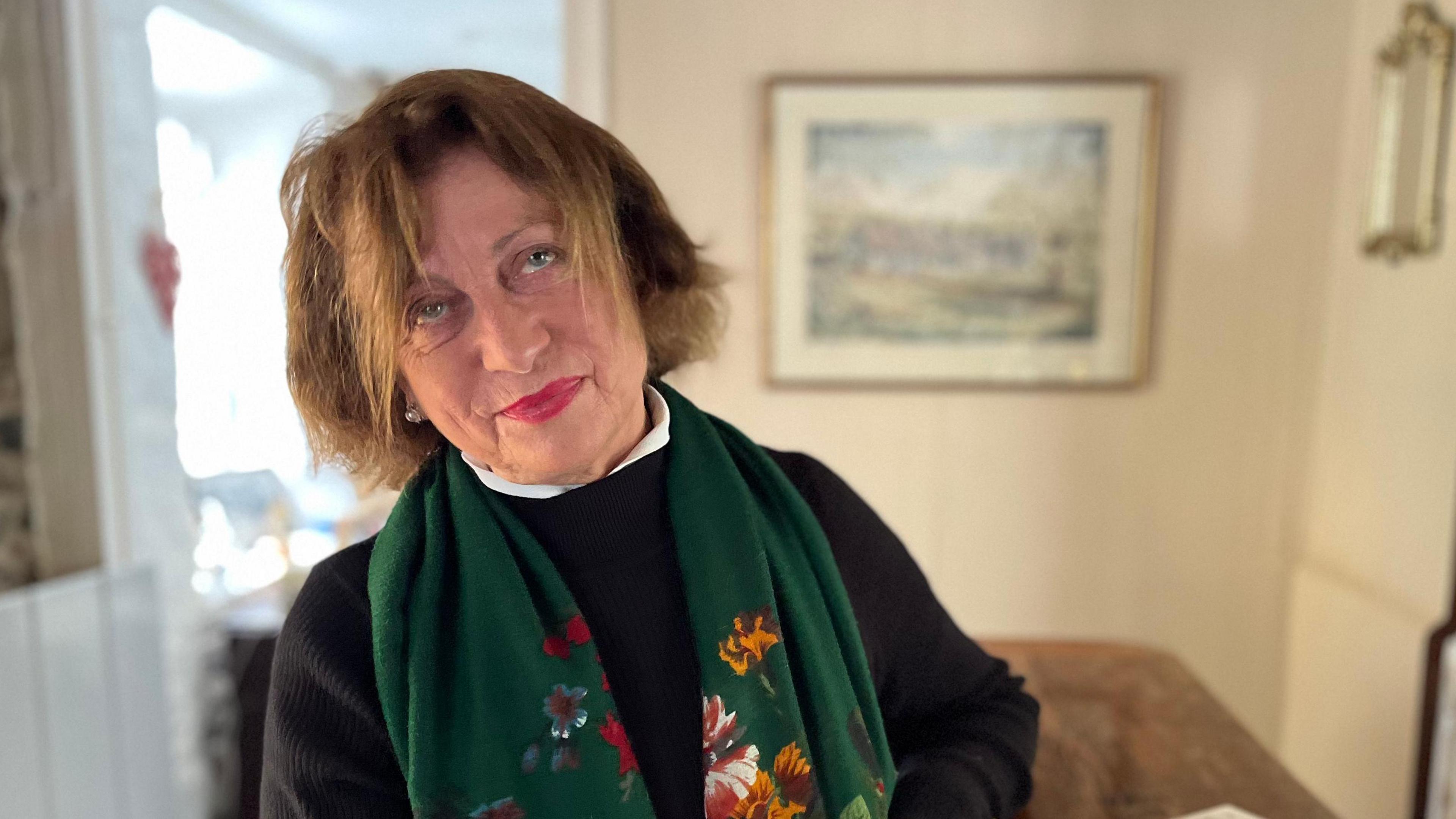 Elizabeth Perrée - A woman with brown hair and red lipstick tilting her head and looking at the camera. She's wearing a navy blue dress and a green patterned scarf. 