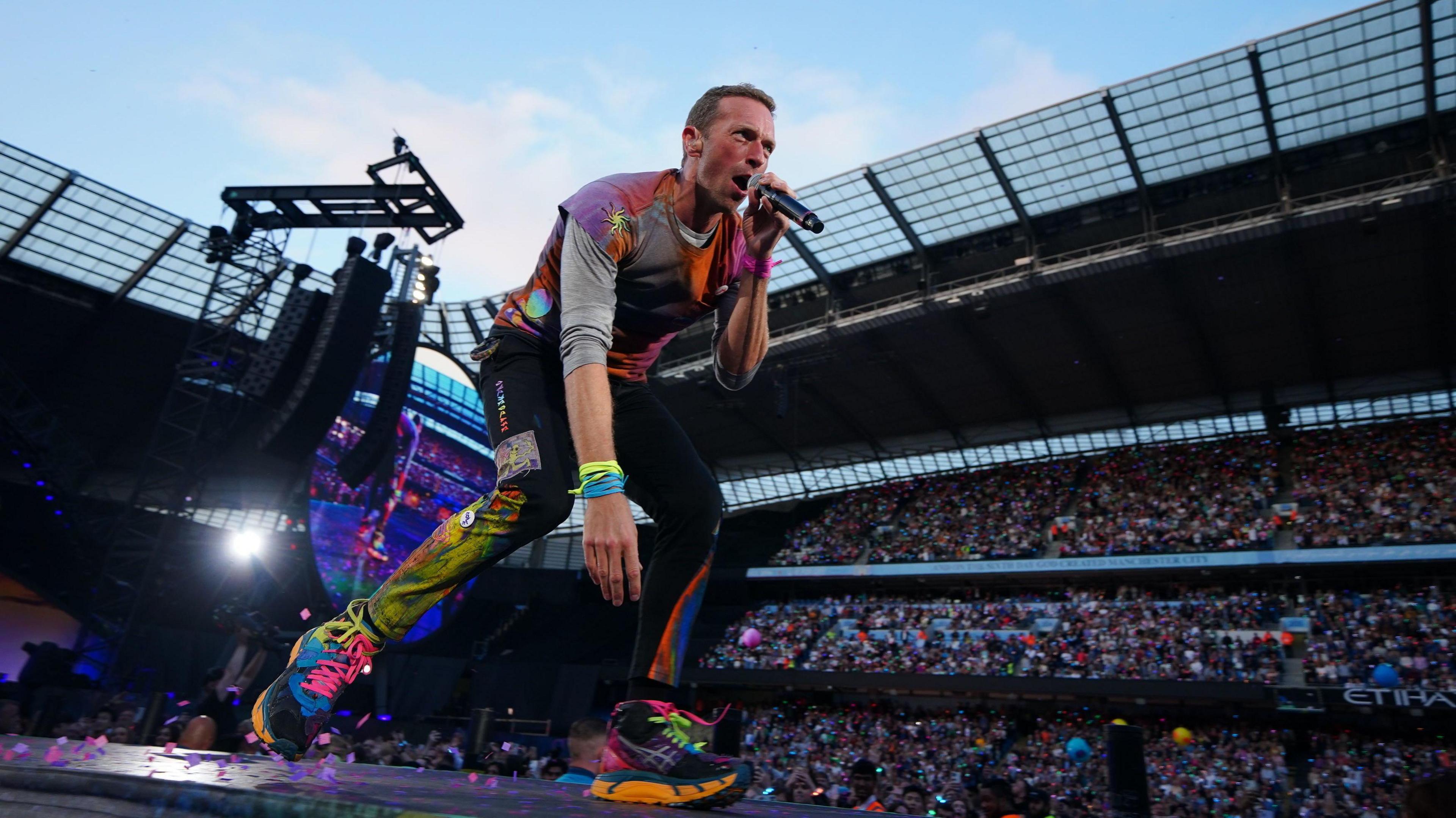 Coldplay lead singer Chris Martin on stage, wearing fluorescent clothing is bending down and singing. Behind him are thousands of fans in a stadium